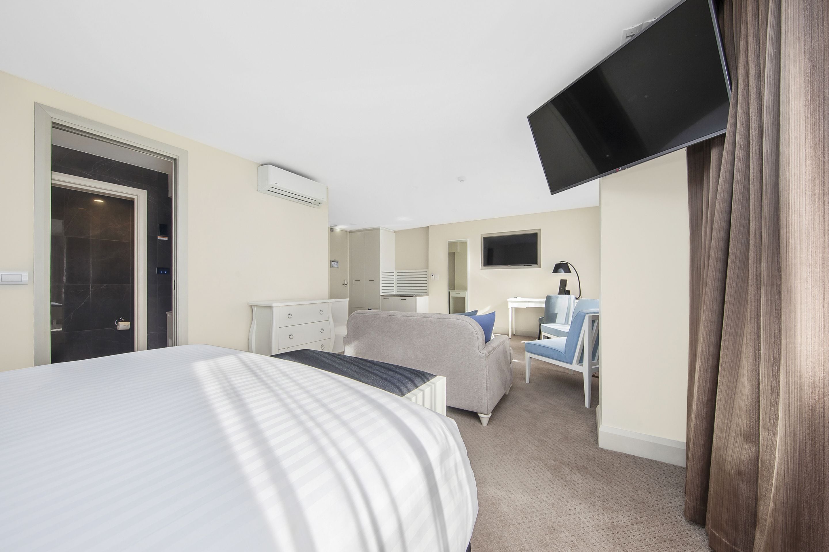 Habitación de hotel con una cama grande, un escritorio, una silla, un televisor instalado en la pared y un baño visible a través de una puerta abierta.