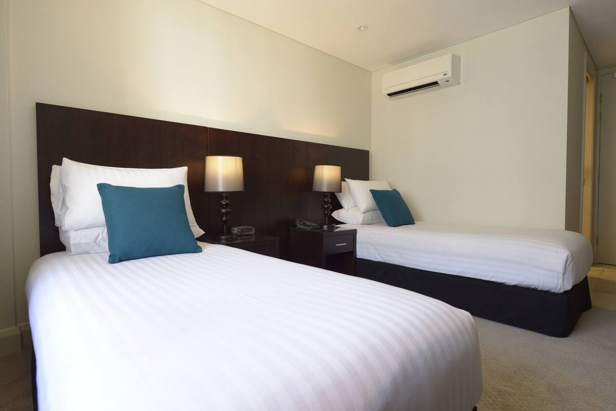 A hotel room with two beds, each with white linens and teal pillows, a dark headboard, bedside tables with lamps, and an air conditioning unit on the wall.