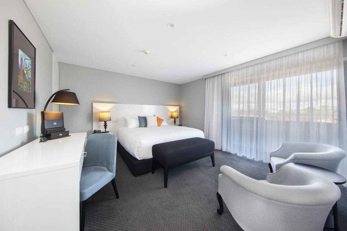 A hotel room with a bed, two bedside tables, a desk, a chair, an ottoman, and a large window with sheer curtains.