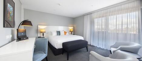Habitación de hotel con cama, dos mesitas de noche, escritorio, silla, reposapiés y una ventana grande con cortinas translúcidas.