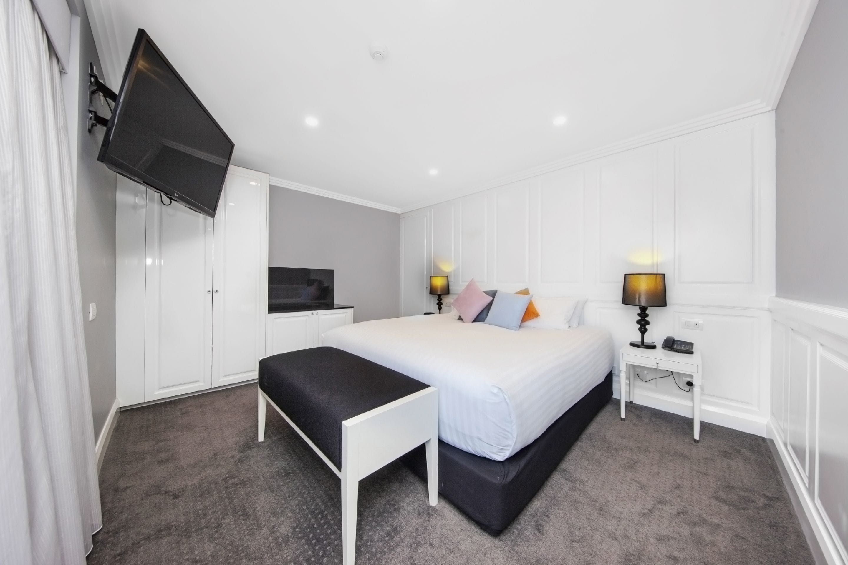 Une chambre à coucher avec un lit, des tables de chevet, un téléviseur fixé au mur et un banc.