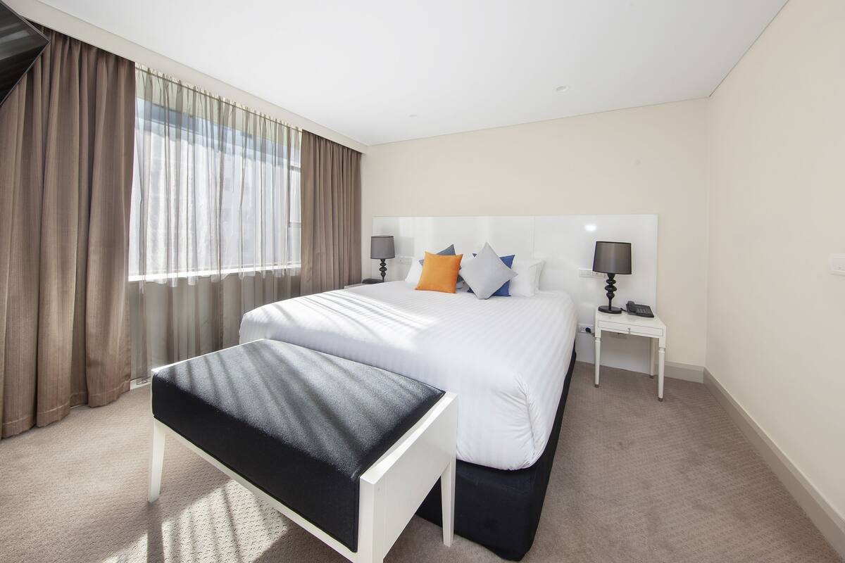 A hotel room with a large bed, a bedside table with a lamp, a bench, and a window with curtains.