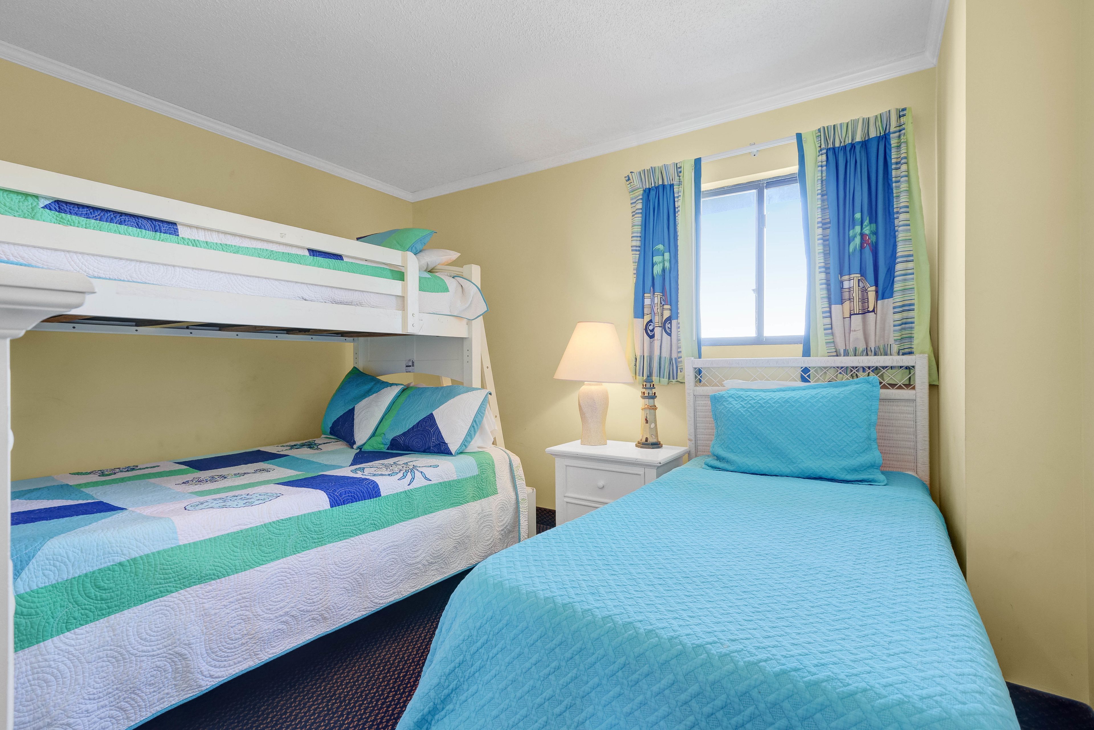 Quarto de hotel com uma cama beliche, uma cama de solteiro, uma mesa de cabeceira com luminária e uma janela com cortinas.