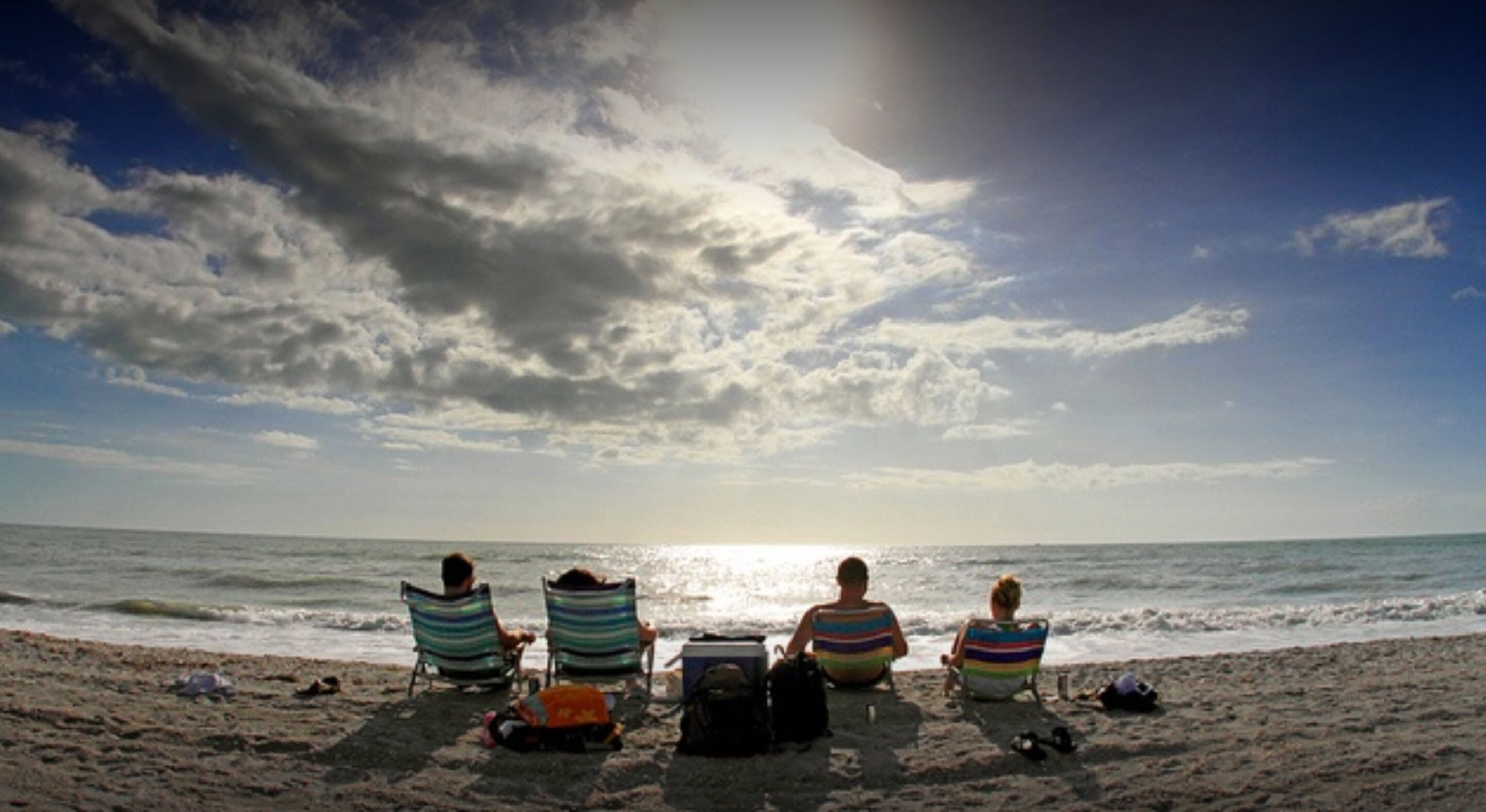 Une plage où des personnes sont assises sur des chaises, face à l’océan, sous un ciel partiellement nuageux.
