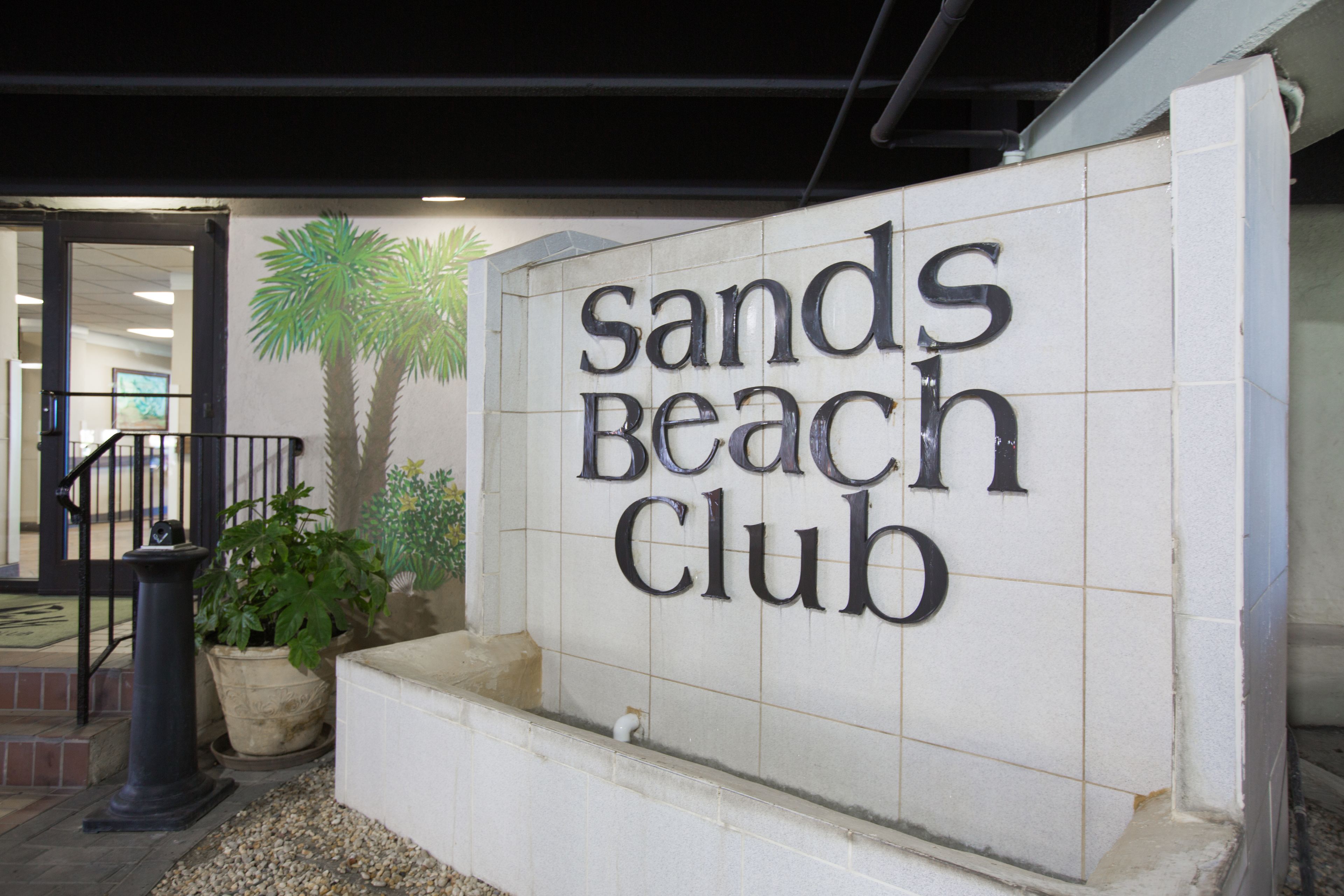 A sign for the Sands Beach Club with a tiled wall and a palm tree.