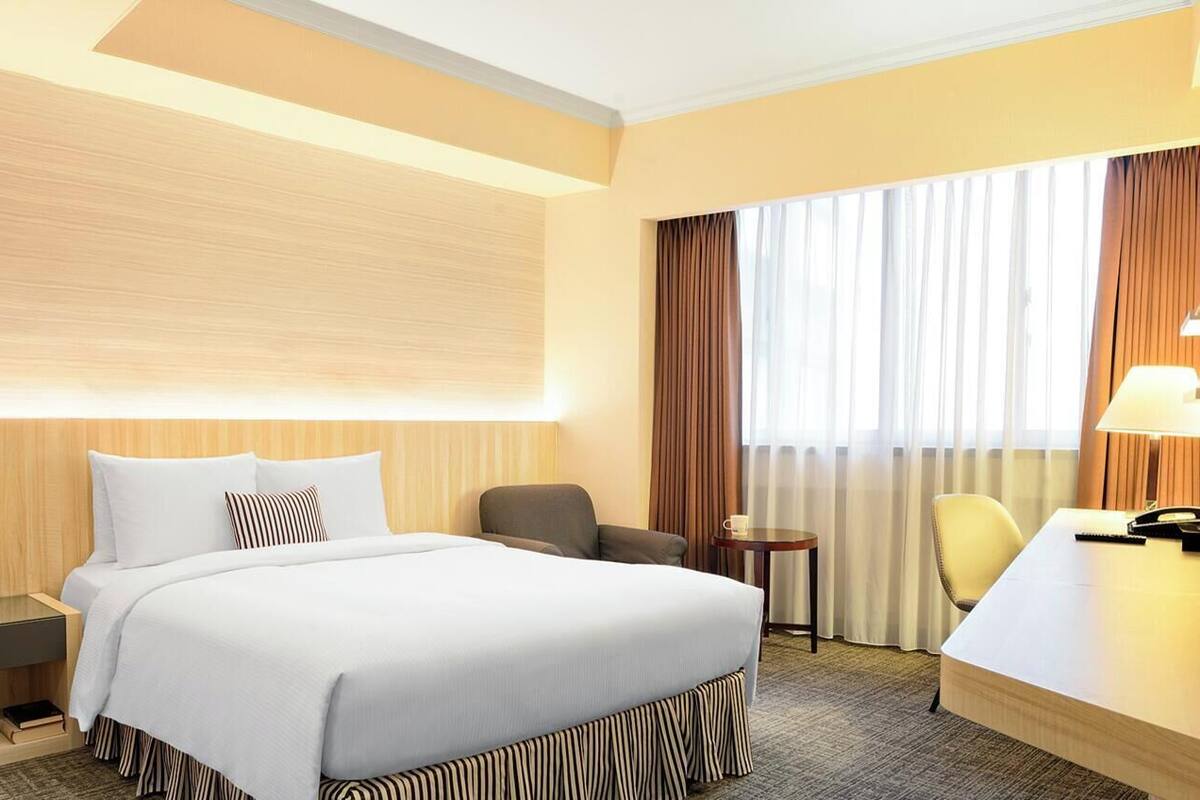 A hotel room with a bed, bedside table, lamp, chair, and a desk with a telephone.