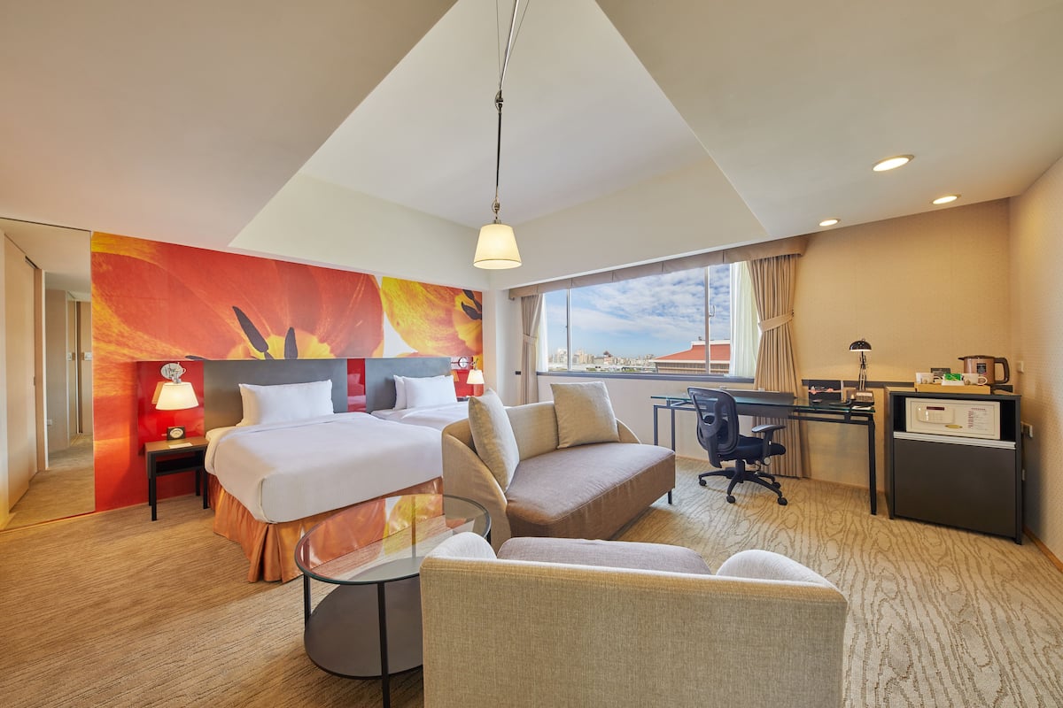A hotel room with a bed, sofa, desk, and chair. There is a window with a view of buildings, a lamp, and a clock on the nightstand.