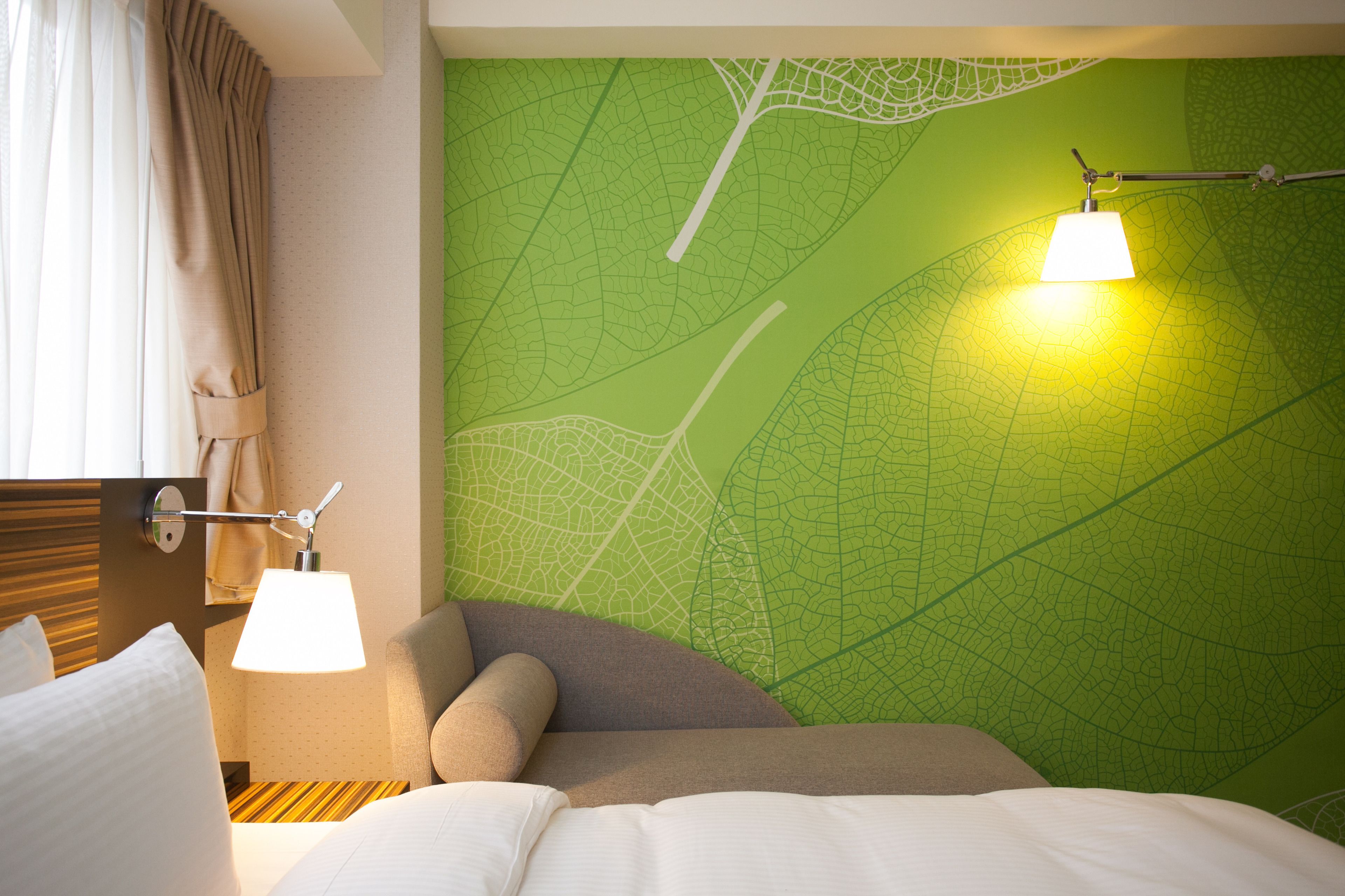 Una habitación de hotel con un papel tapiz de hojas verdes, una cama con ropa blanca, un sillón marrón y dos lámparas de noche.