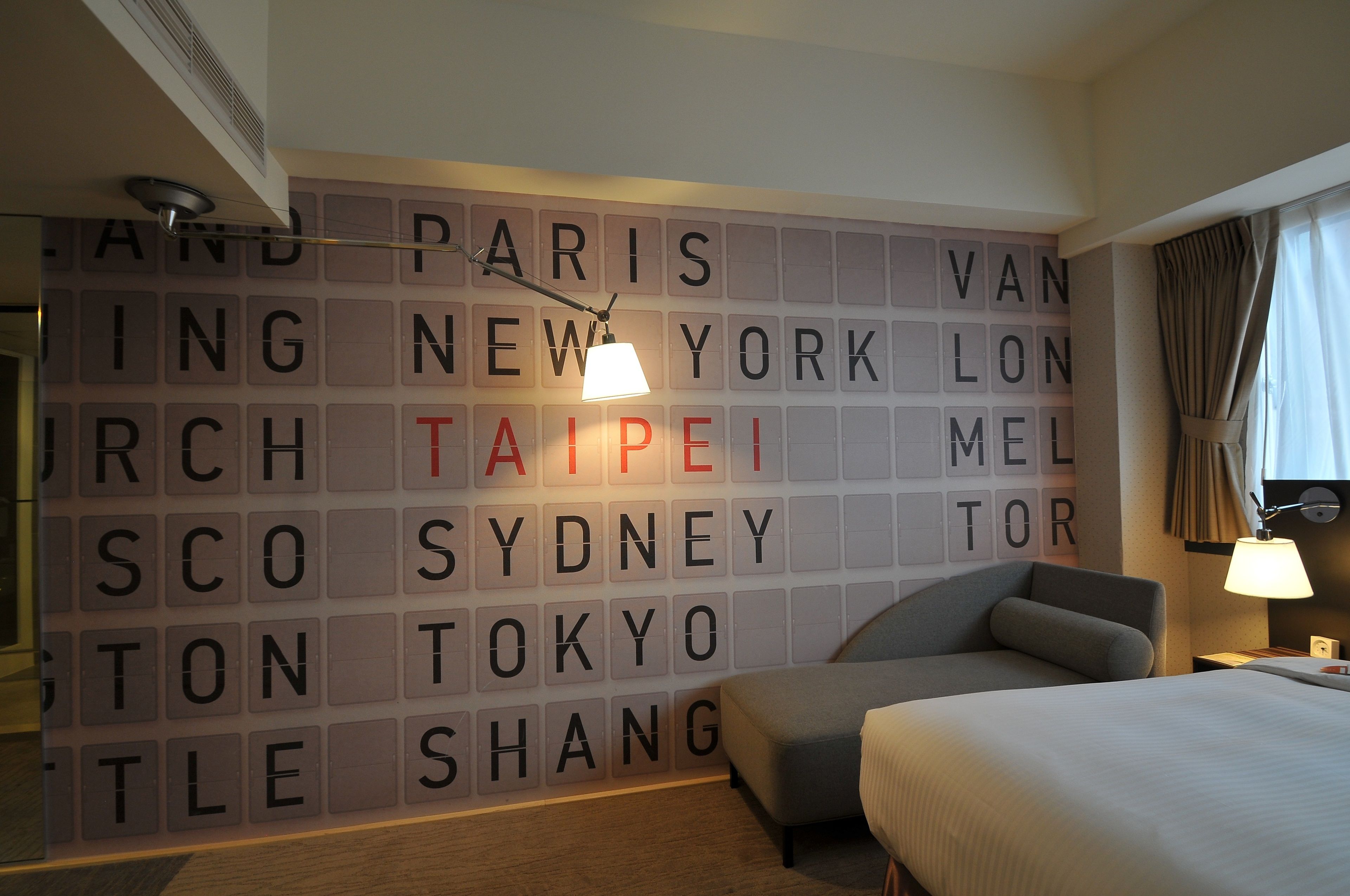 A hotel room with a wall displaying city names in large letters, a bed, a chair, and a nightstand with a lamp.