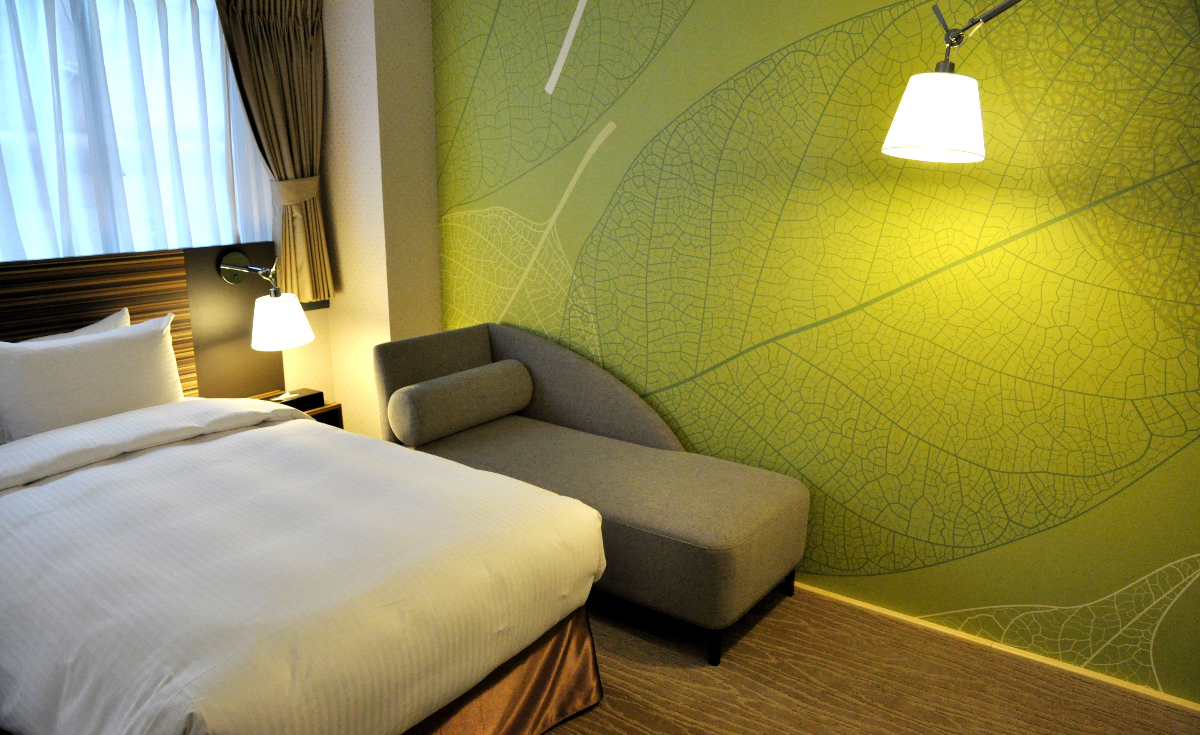 Una habitación de hotel con una cama, una mesita de noche, una lámpara, una silla y una pared con estampado de hojas verdes.