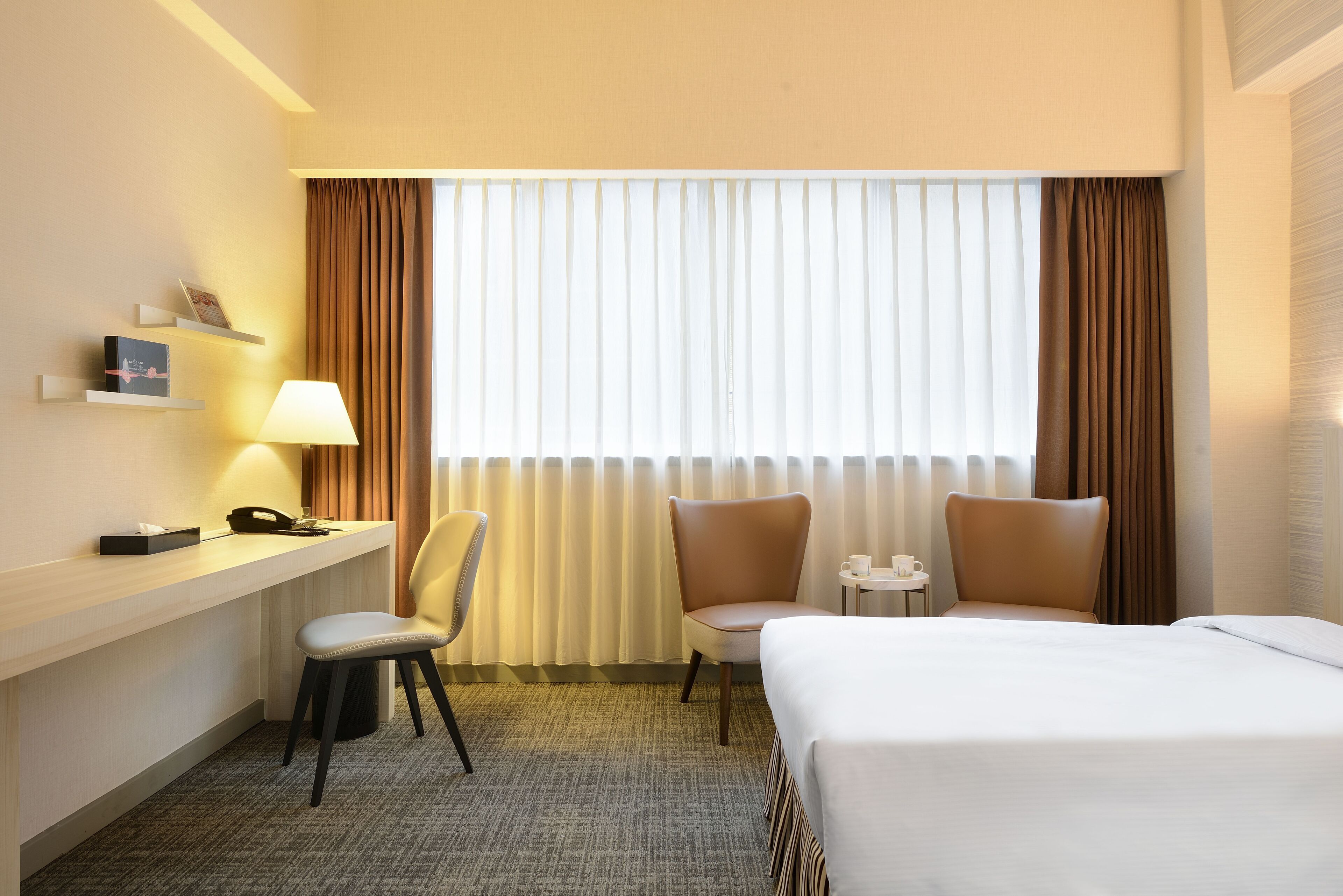 Habitación de hotel con una cama, dos sillas, un escritorio con lámpara y una ventana con cortinas.