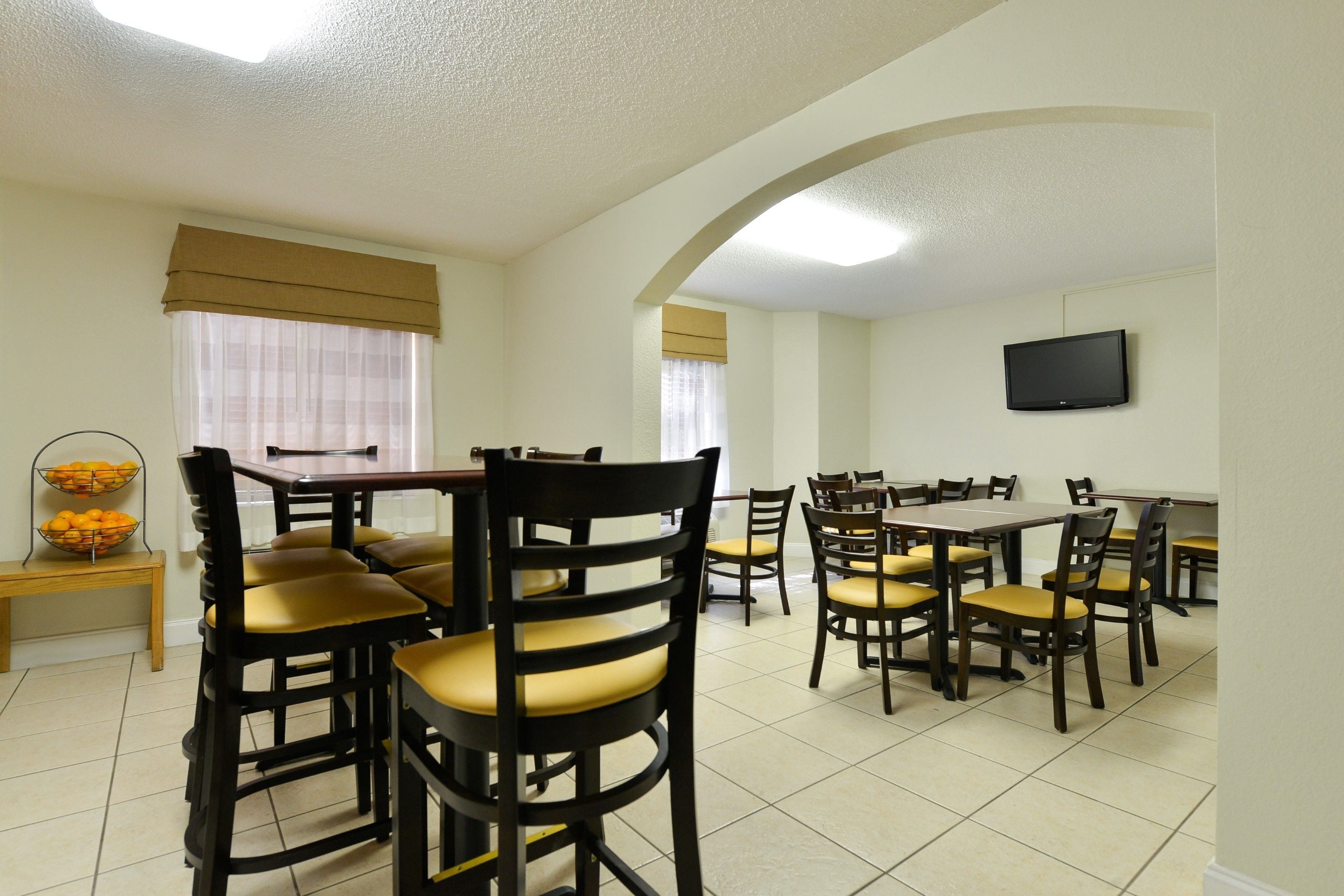 A dining area with multiple tables and chairs, a television mounted on the wall, and a window with curtains.
