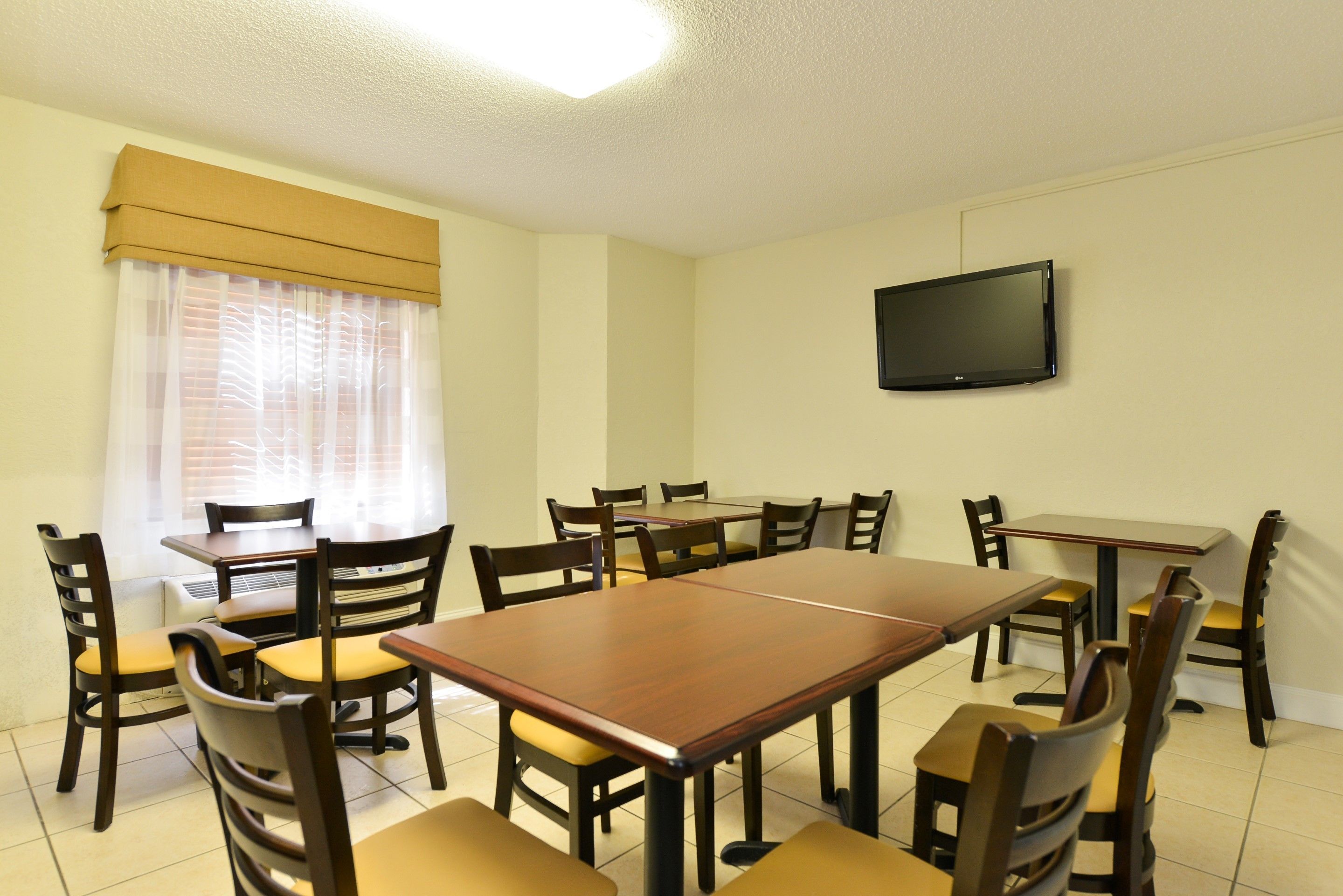 A dining area with a large wooden table and multiple chairs, a television mounted on the wall, and a window with sheer curtains.