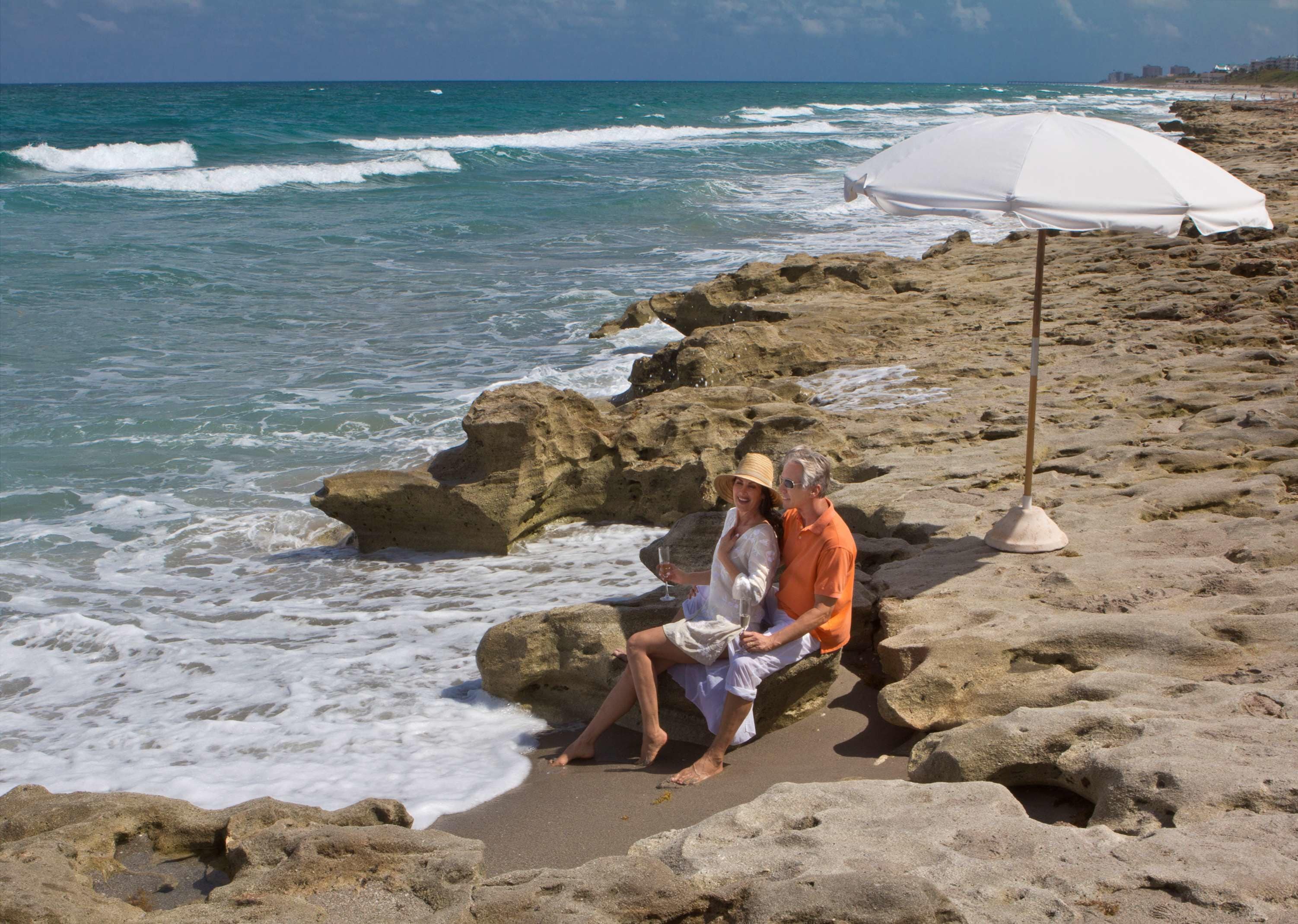 A couple sitting on a rocky beach with a white umbrella nearby.