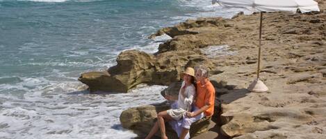 A couple sitting on a rocky beach with a white umbrella nearby.