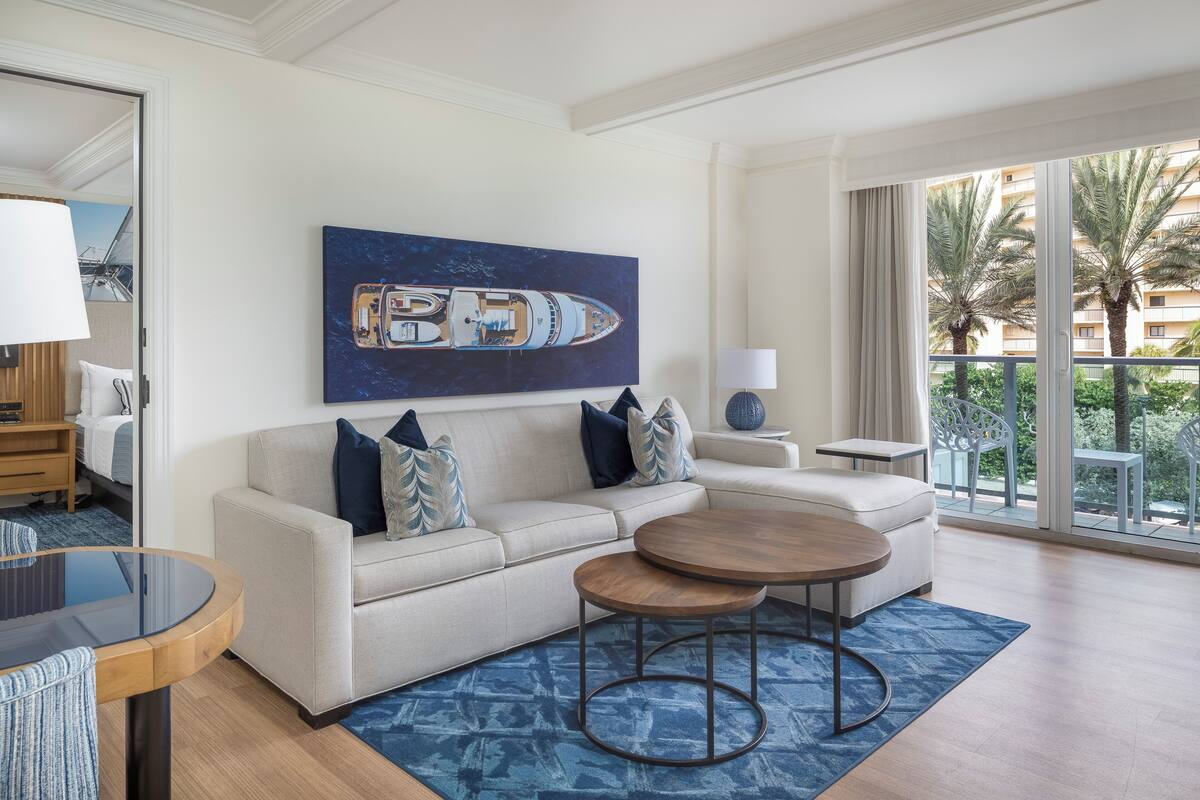A modern living room with a sofa, two round tables, a large painting of a yacht, and a balcony with palm trees.