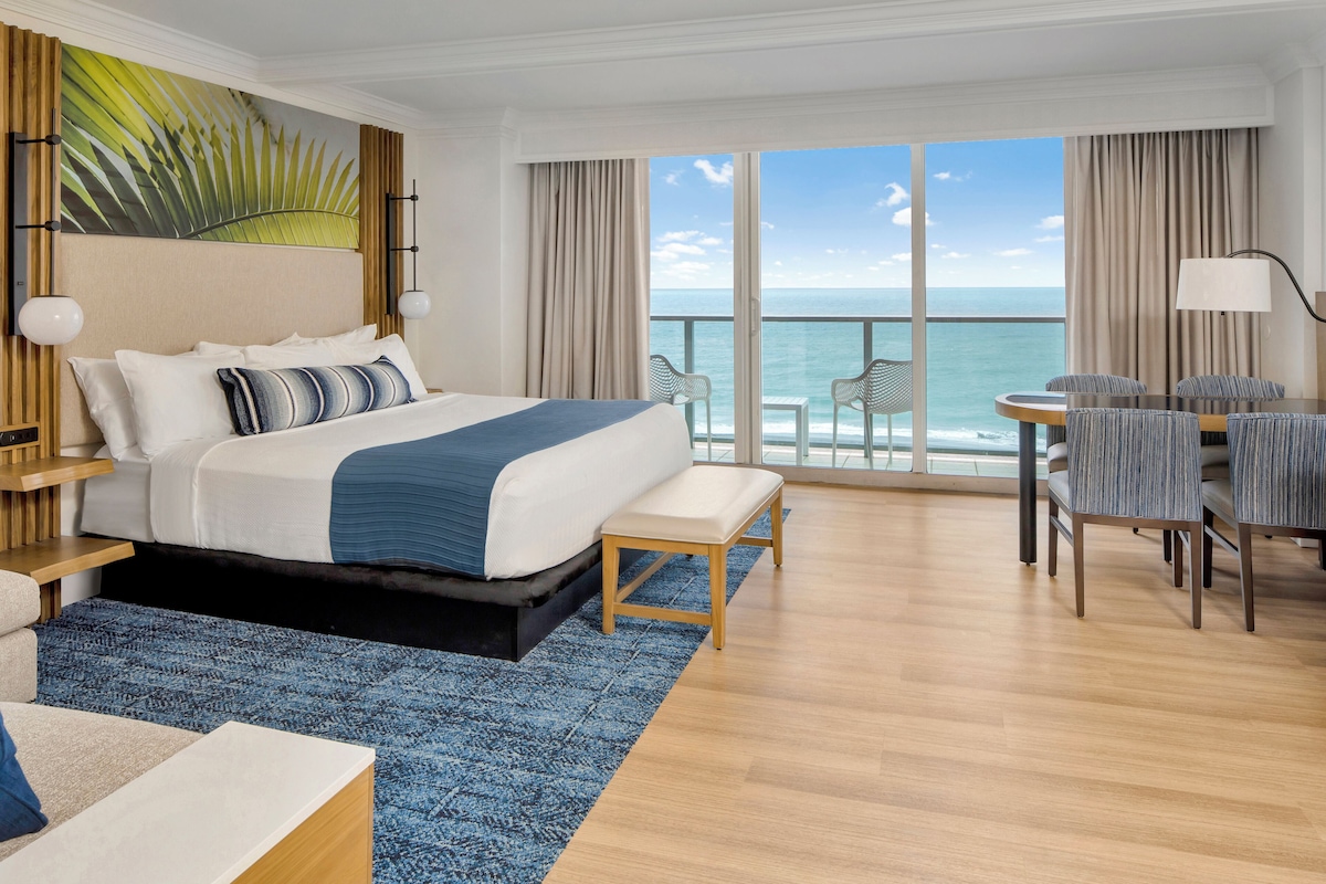 A hotel room with a large bed, a balcony with ocean views, a dining area with chairs, and a large window.