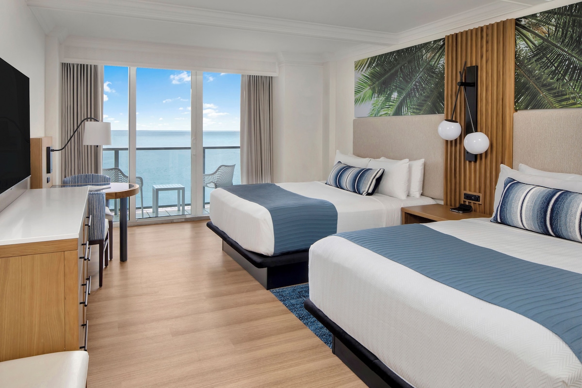 A hotel room with two beds, a desk, and a chair. There is a large window with a view of the ocean.