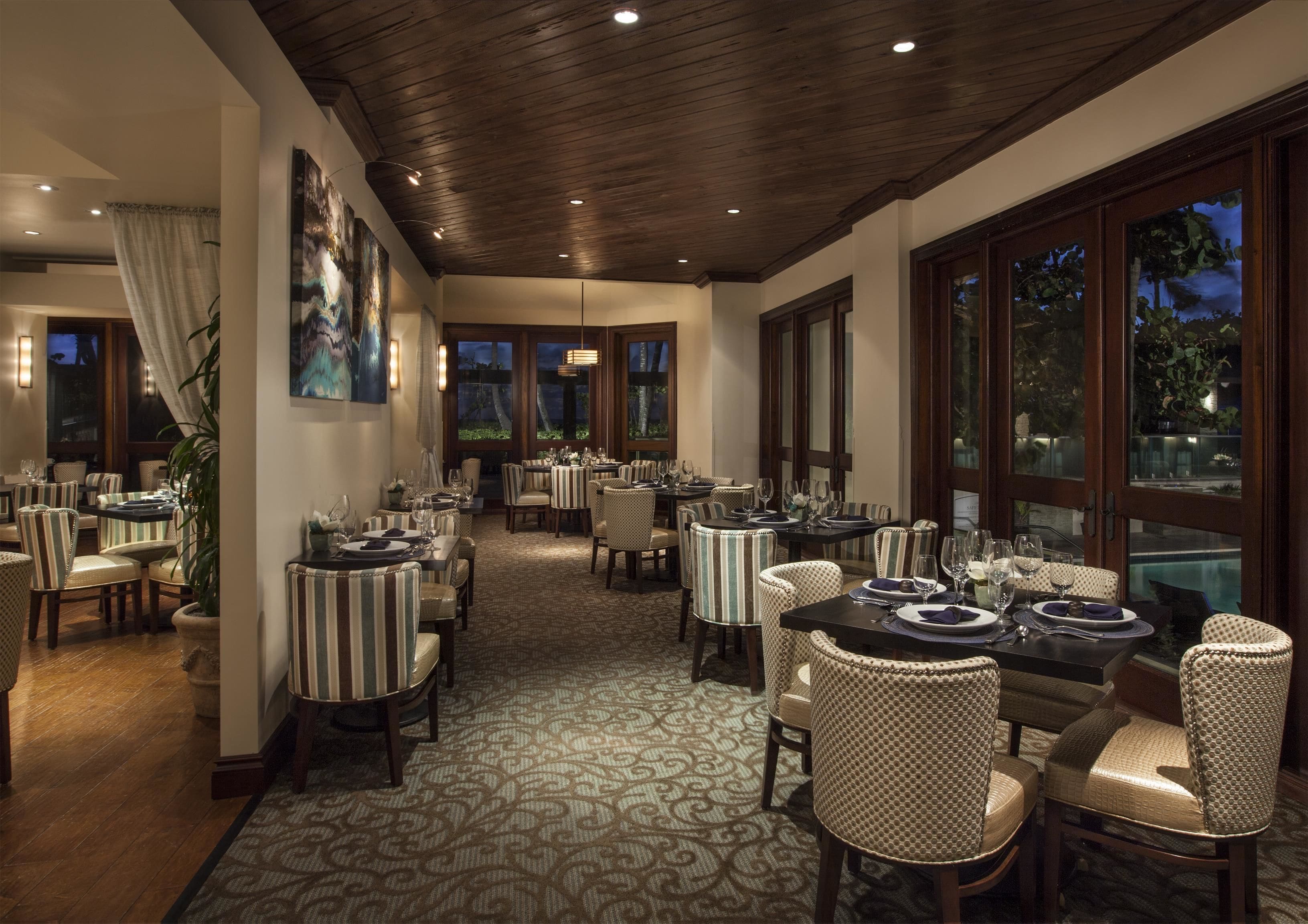 A restaurant with multiple tables and chairs, large windows, and a patterned carpet.