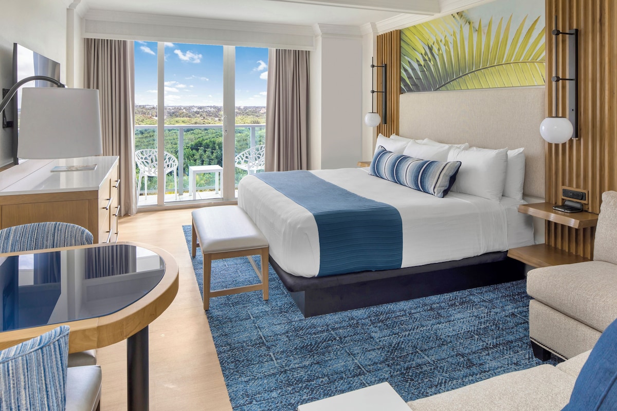 A hotel room with a large bed, a desk with a chair, a small table, and a balcony with a view.