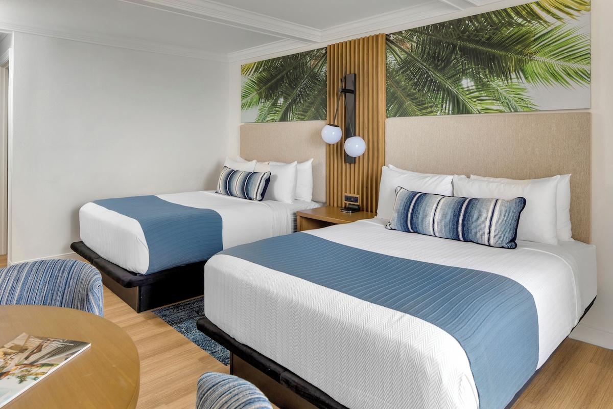 A hotel room with two beds, a wooden headboard, a nightstand, a lamp, and a view of palm trees through a window.