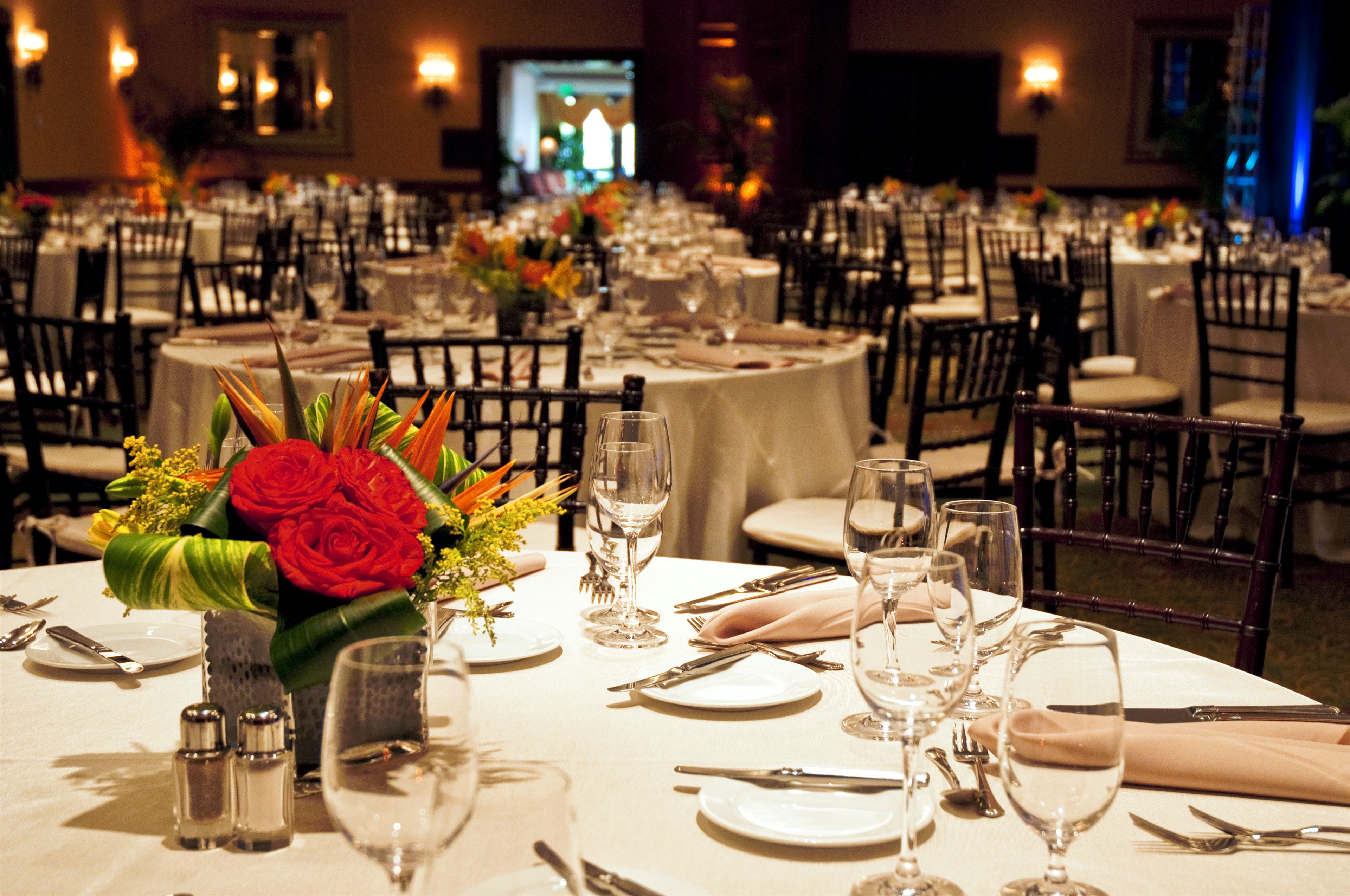 Un salón de banquetes con mesas redondas dispuestas para cenar, incluyendo vajilla, sillas y centros de mesa con flores.