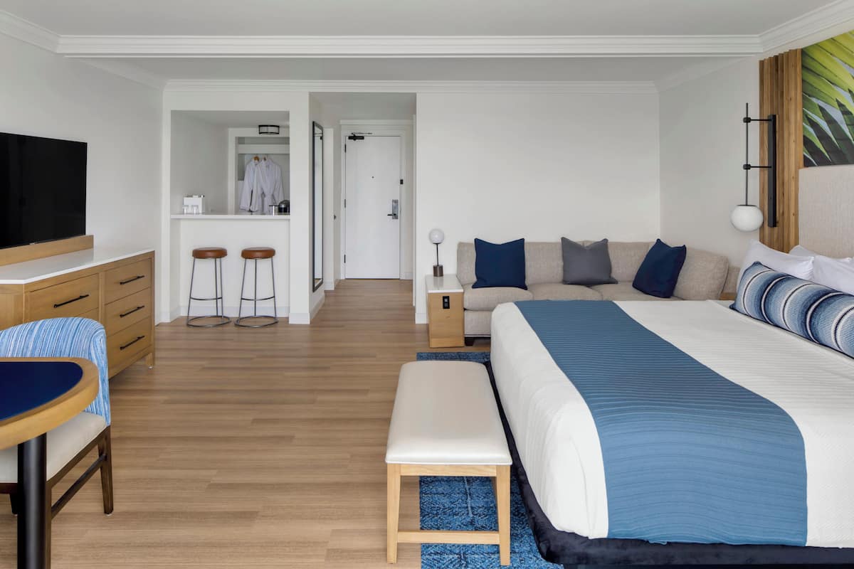 A modern hotel room with a large bed, a desk with a chair, a television, and a small kitchenette area with stools.