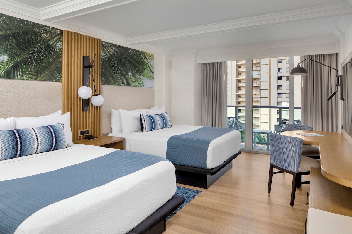 A hotel room with two beds, a desk, and a view of palm trees and buildings.