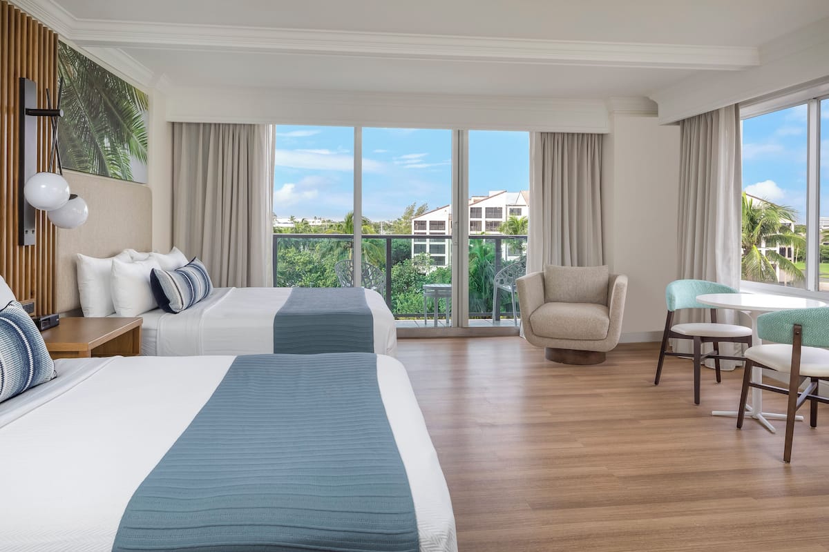 A hotel room with two beds, a dining area, and large windows with a view of palm trees and buildings.