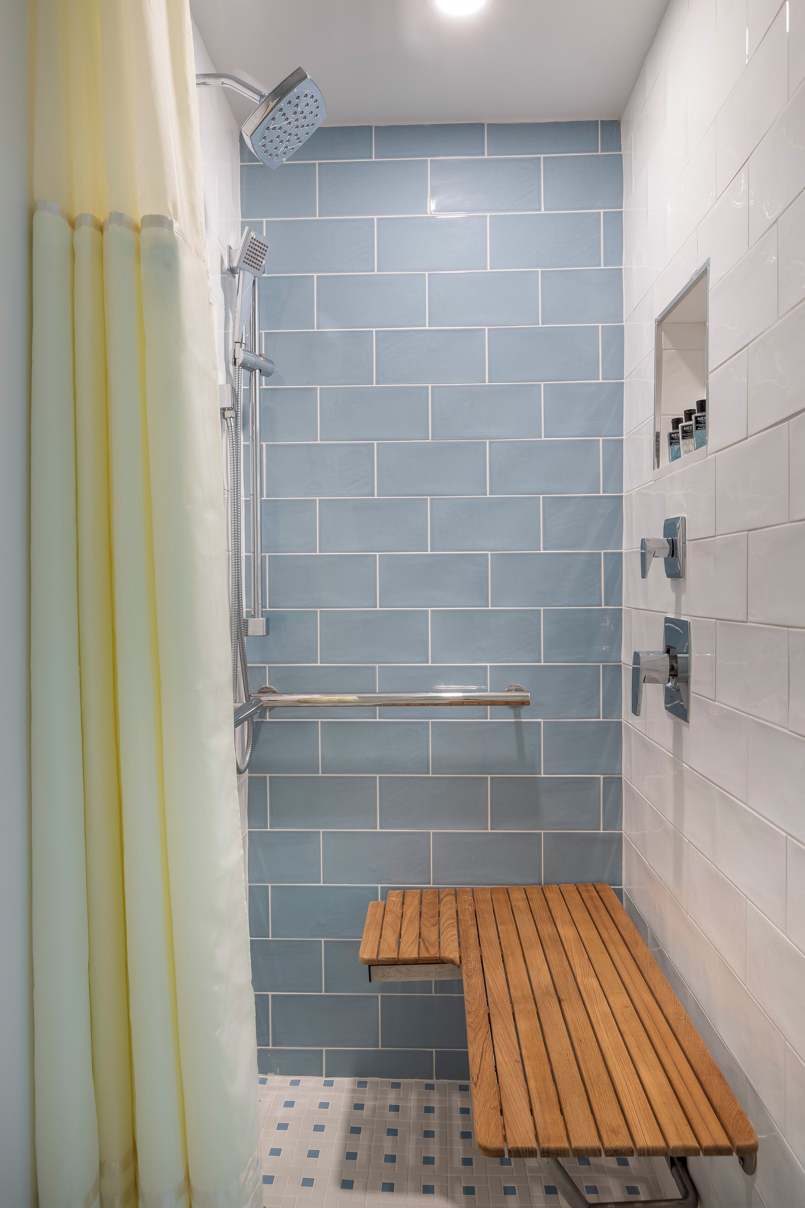 A shower with a handheld showerhead, a wooden bench, and a tiled wall.