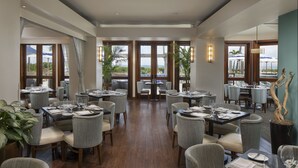 A spacious dining area with multiple tables and chairs, large windows offering views of the ocean, and potted plants. - Jupiter Beach Resort & Spa (Jupiter)