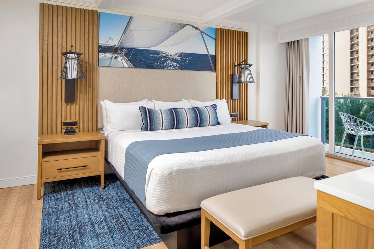 A modern hotel room with a large bed, wooden headboard, and a view of the ocean through the window.