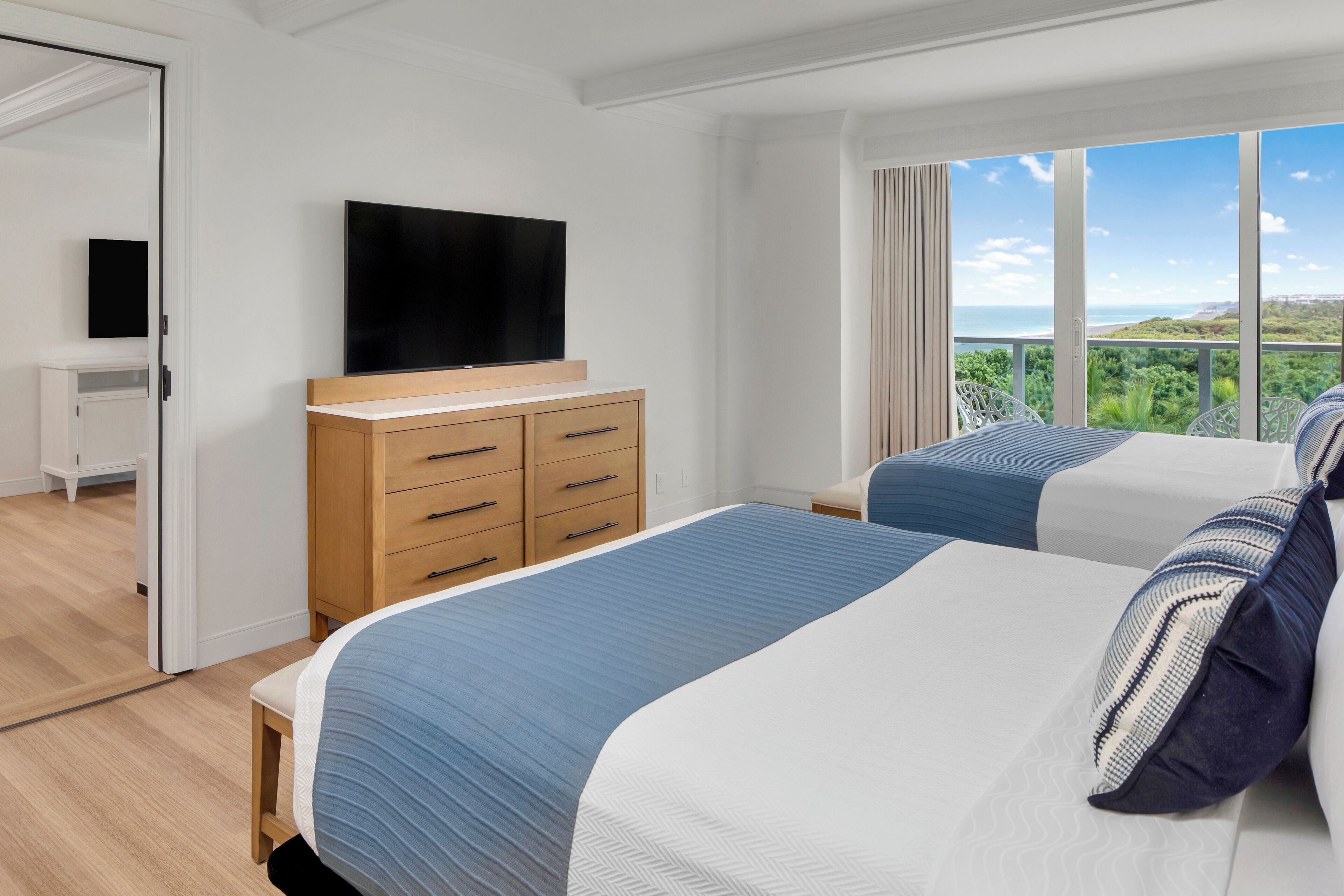 Ein modernes Hotelzimmer mit zwei Betten, einem Flachbildfernseher, einer Holzkommode und einem Balkon mit schöner Aussicht.