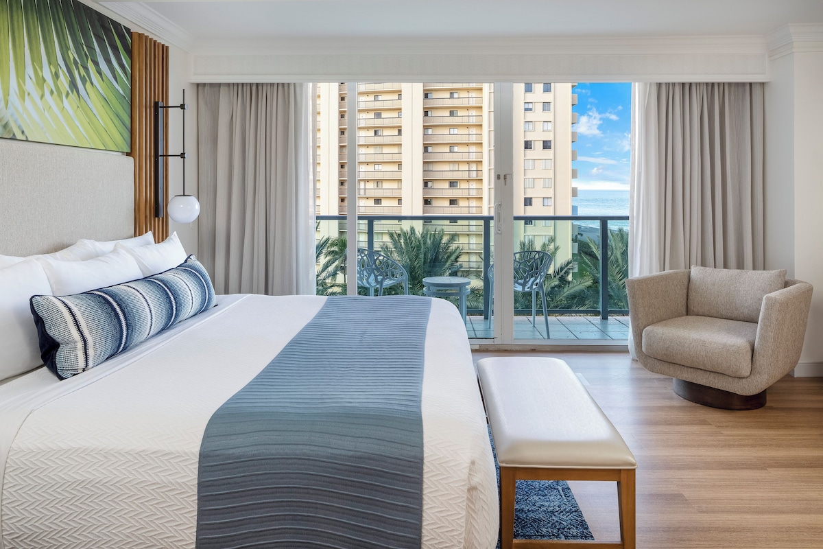 A hotel room with a large bed, a chair, a bench, and a balcony with a view of high-rise buildings and palm trees.
