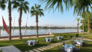An outdoor dining area with tables and chairs, palm trees, and a view of a body of water.