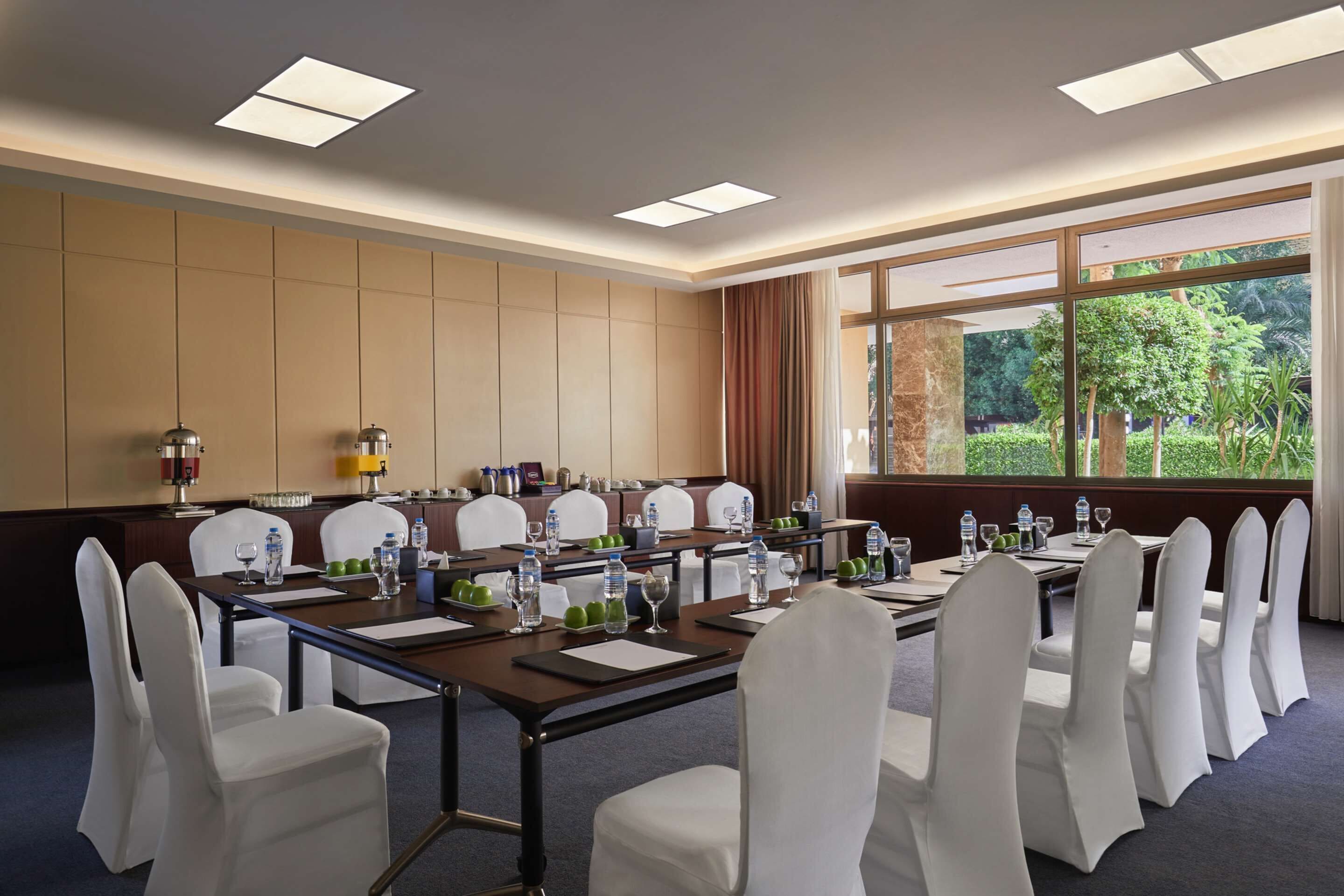 A conference room with a long table, white chairs, and a view of greenery outside.