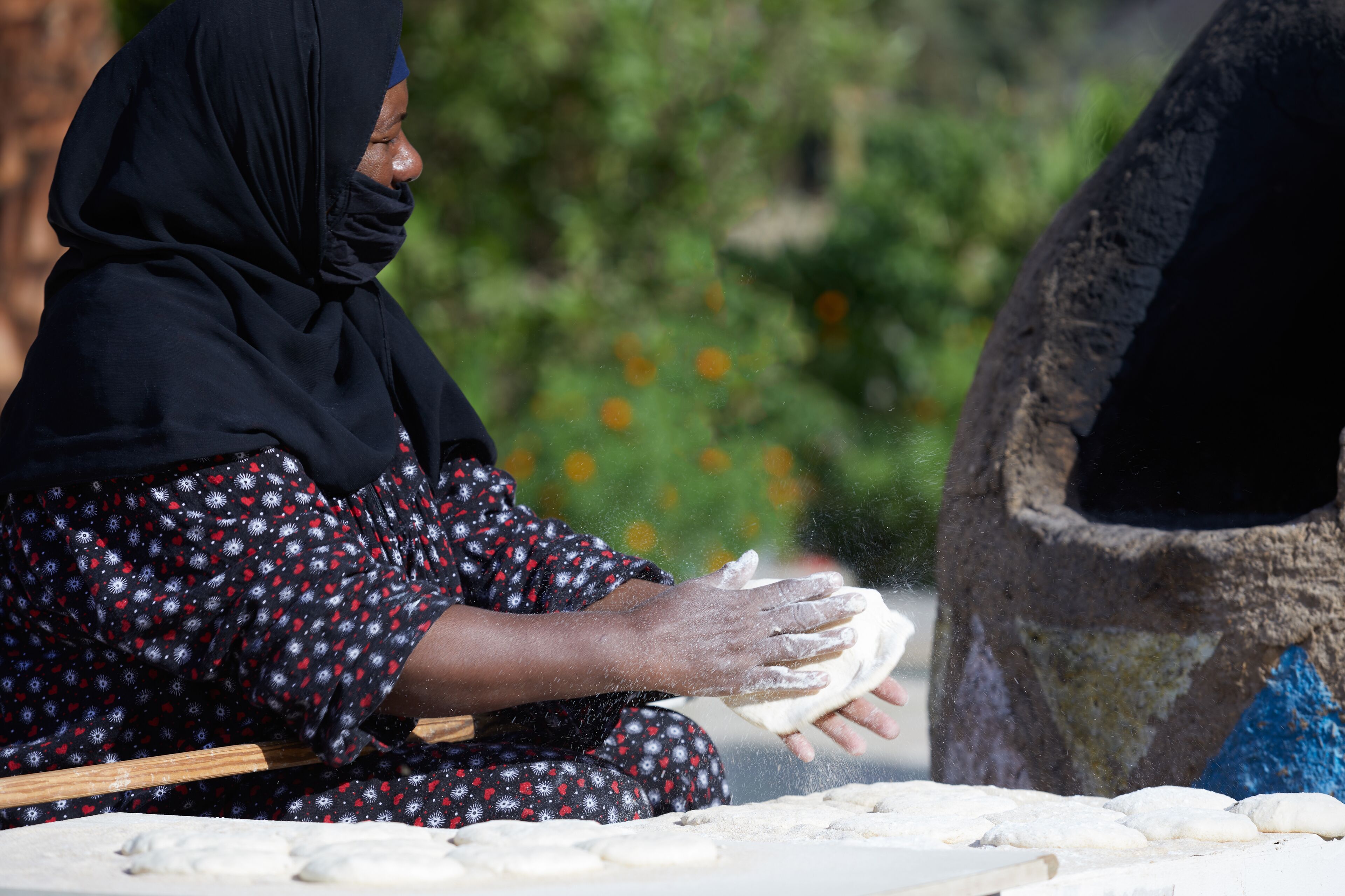 A person kneading dough on a flat surface with a traditional oven in the background.