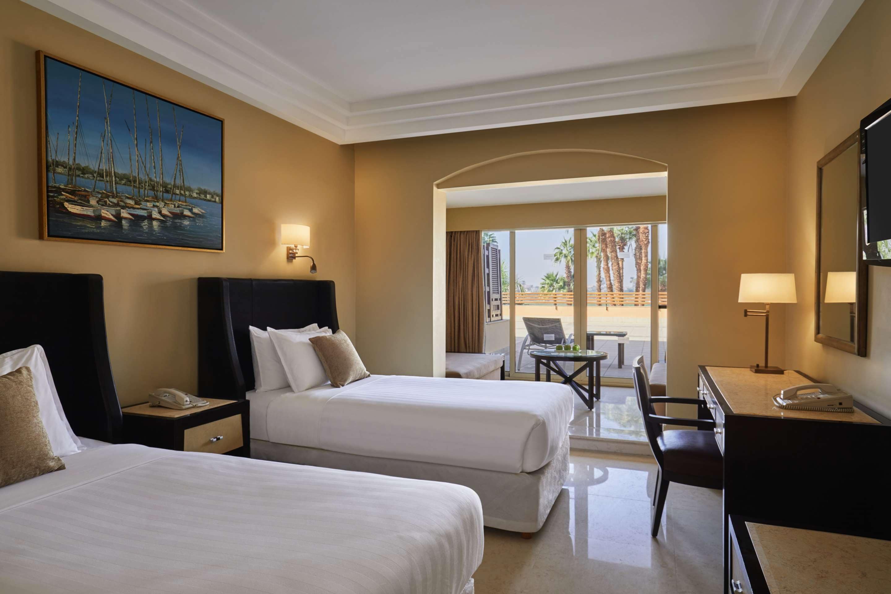 A hotel room with two beds, a desk, a chair, and a painting of sailboats on the wall.