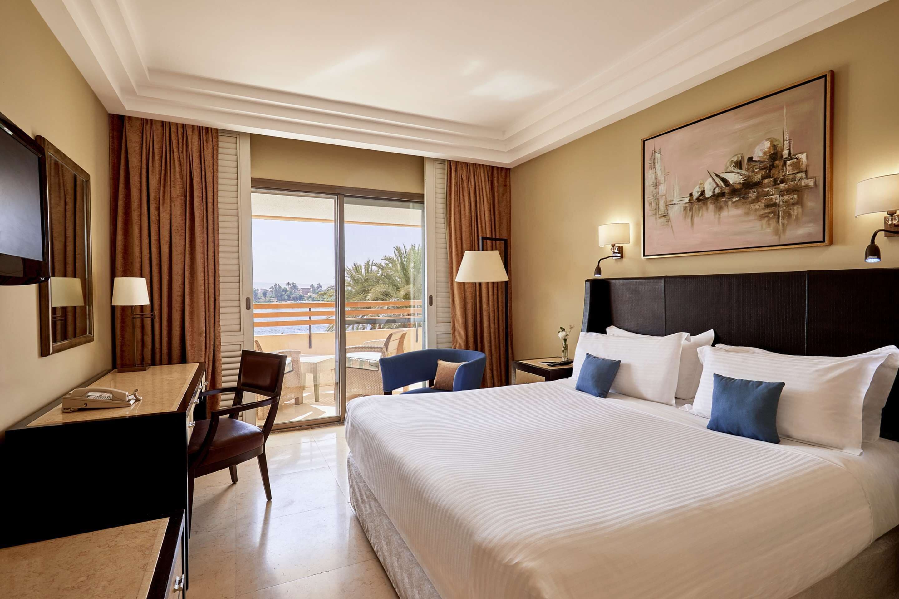A hotel room with a large bed, a desk with a telephone, a chair, a lamp, and a painting on the wall.