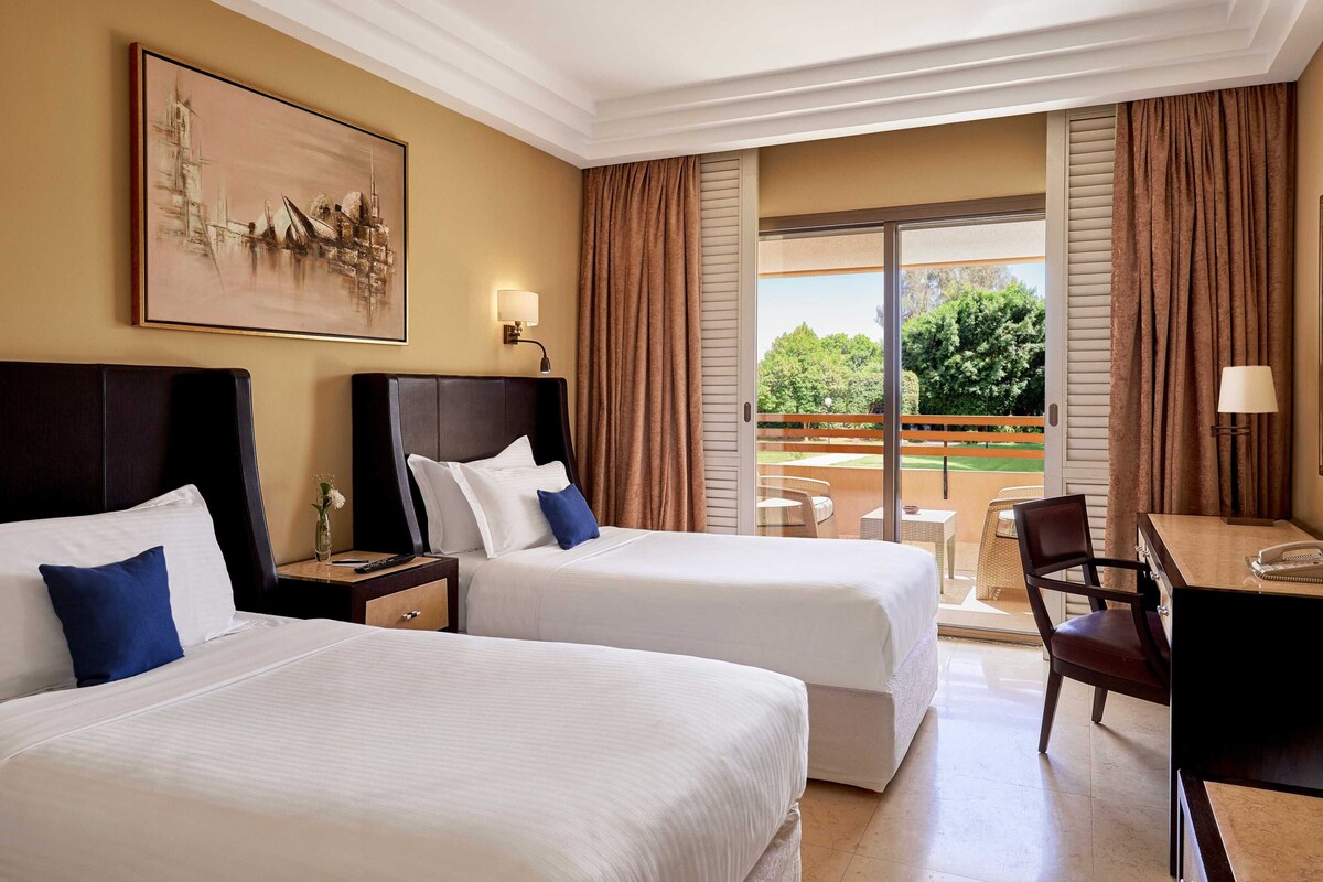 A hotel room with two beds, a desk, a chair, and a balcony with a view of greenery.