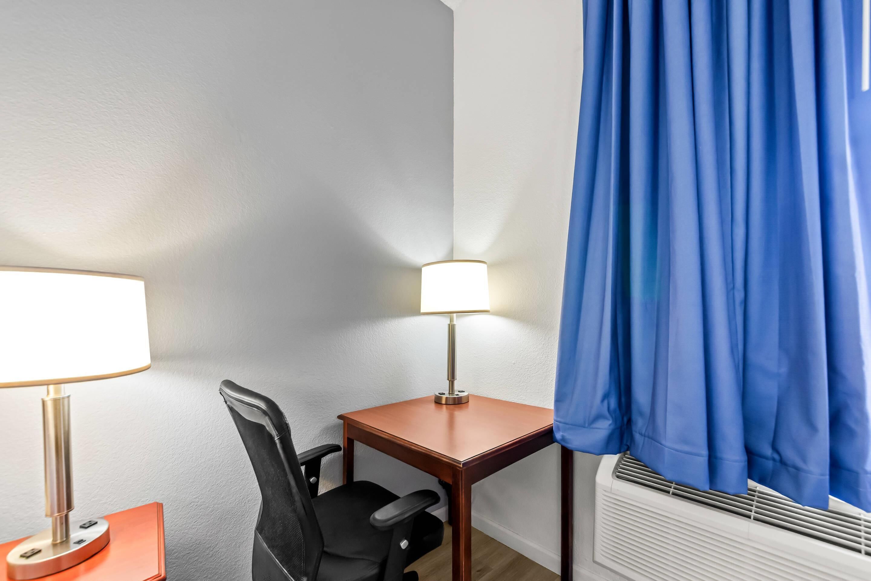 A hotel room with a desk, chair, lamp, and air conditioning unit.