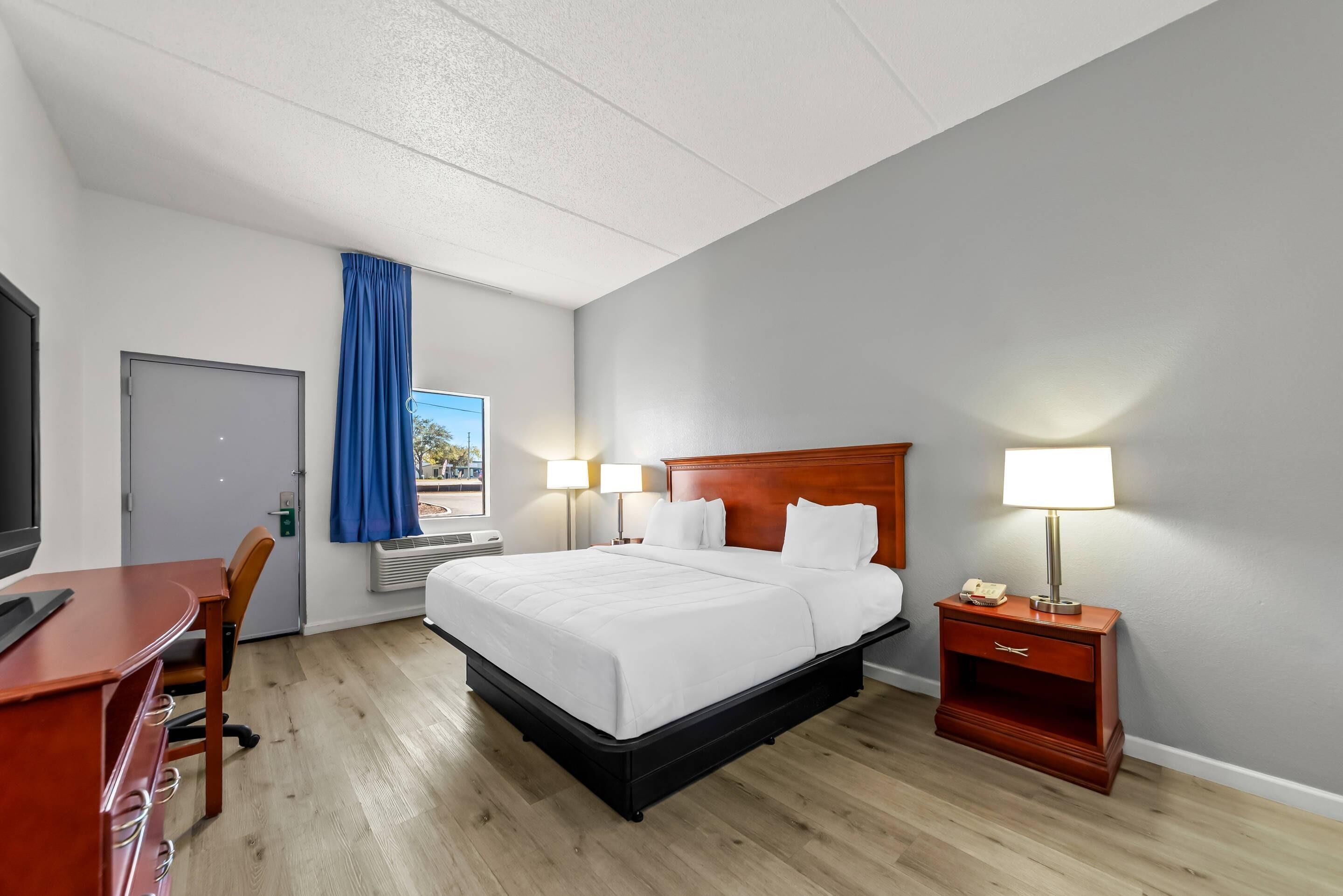 Quarto de hotel com cama, abajures, escrivaninha e televisão.