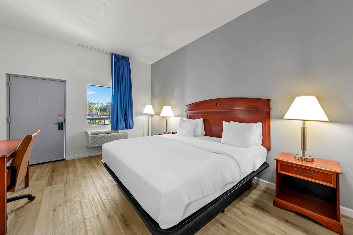 A hotel room with a large bed, two bedside lamps, a wooden nightstand, a desk with a chair, and a window with blue curtains.