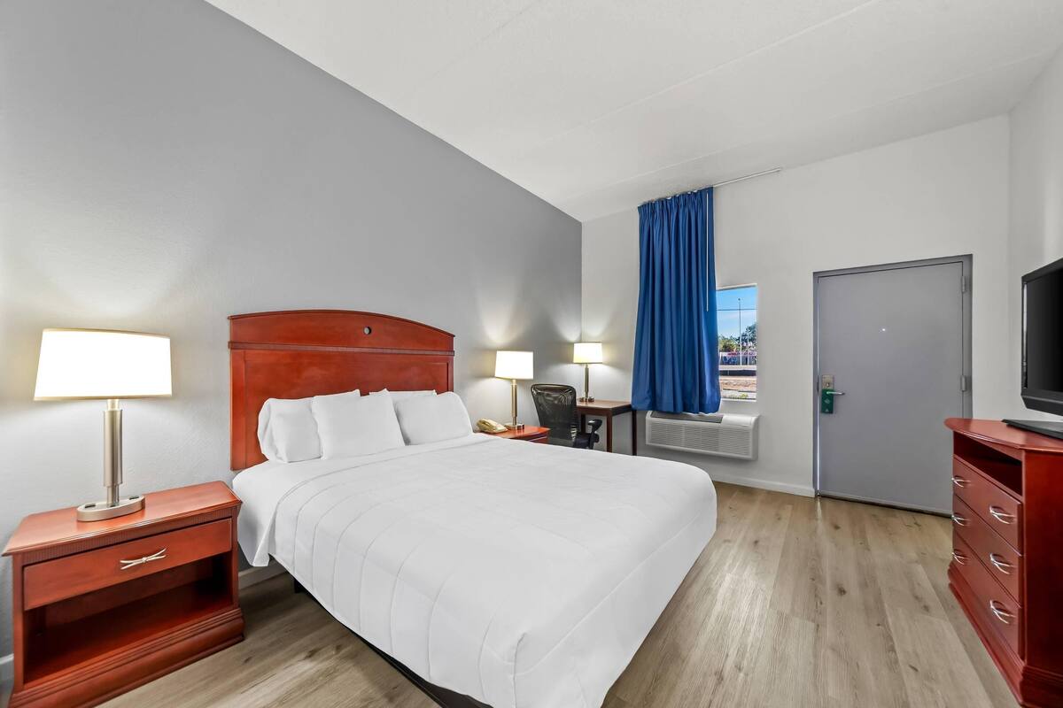 A hotel room with a bed, two bedside tables with lamps, a desk with a chair, a TV, and a wardrobe.