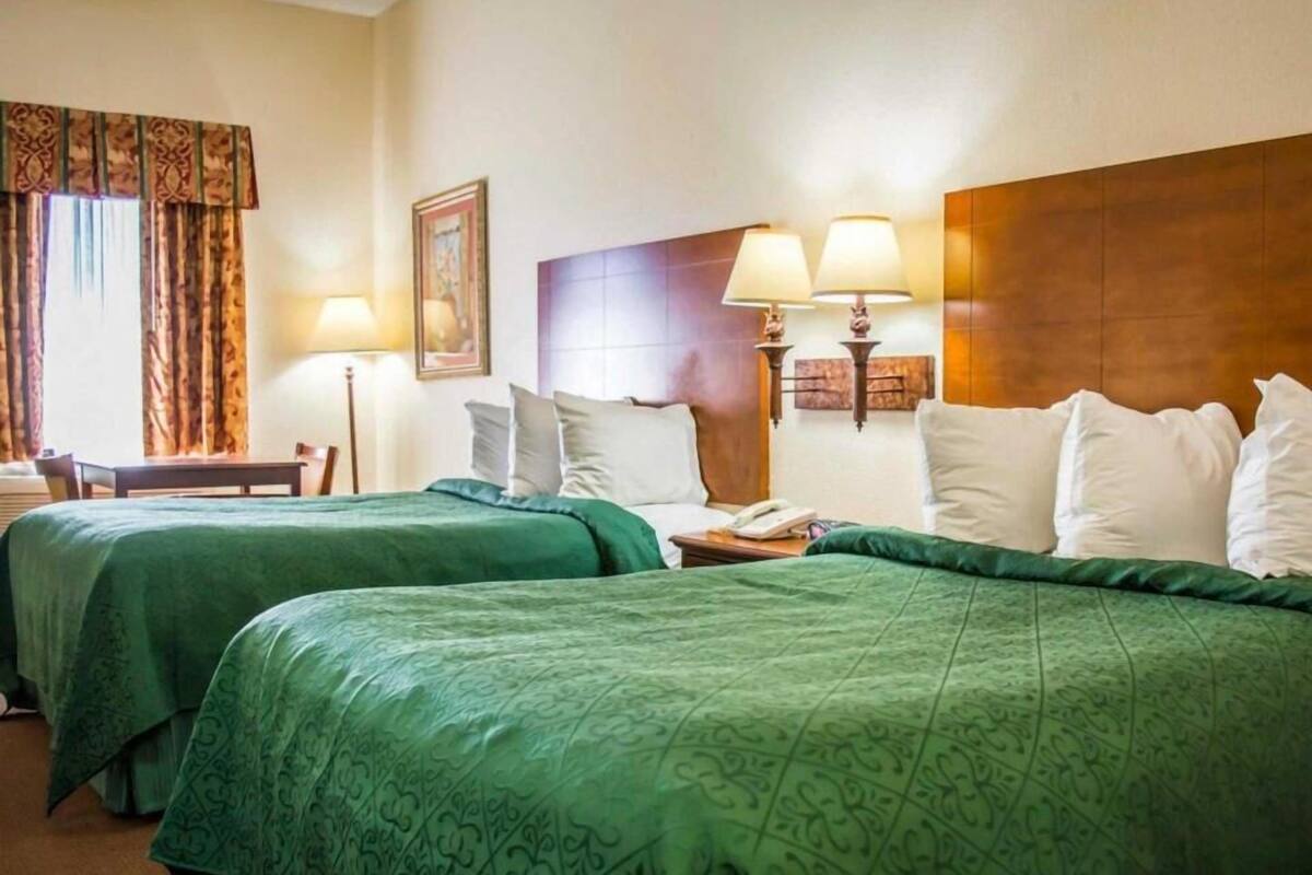 A hotel room with two beds, a wooden headboard, a nightstand with a lamp, a framed picture on the wall, and a window with curtains.