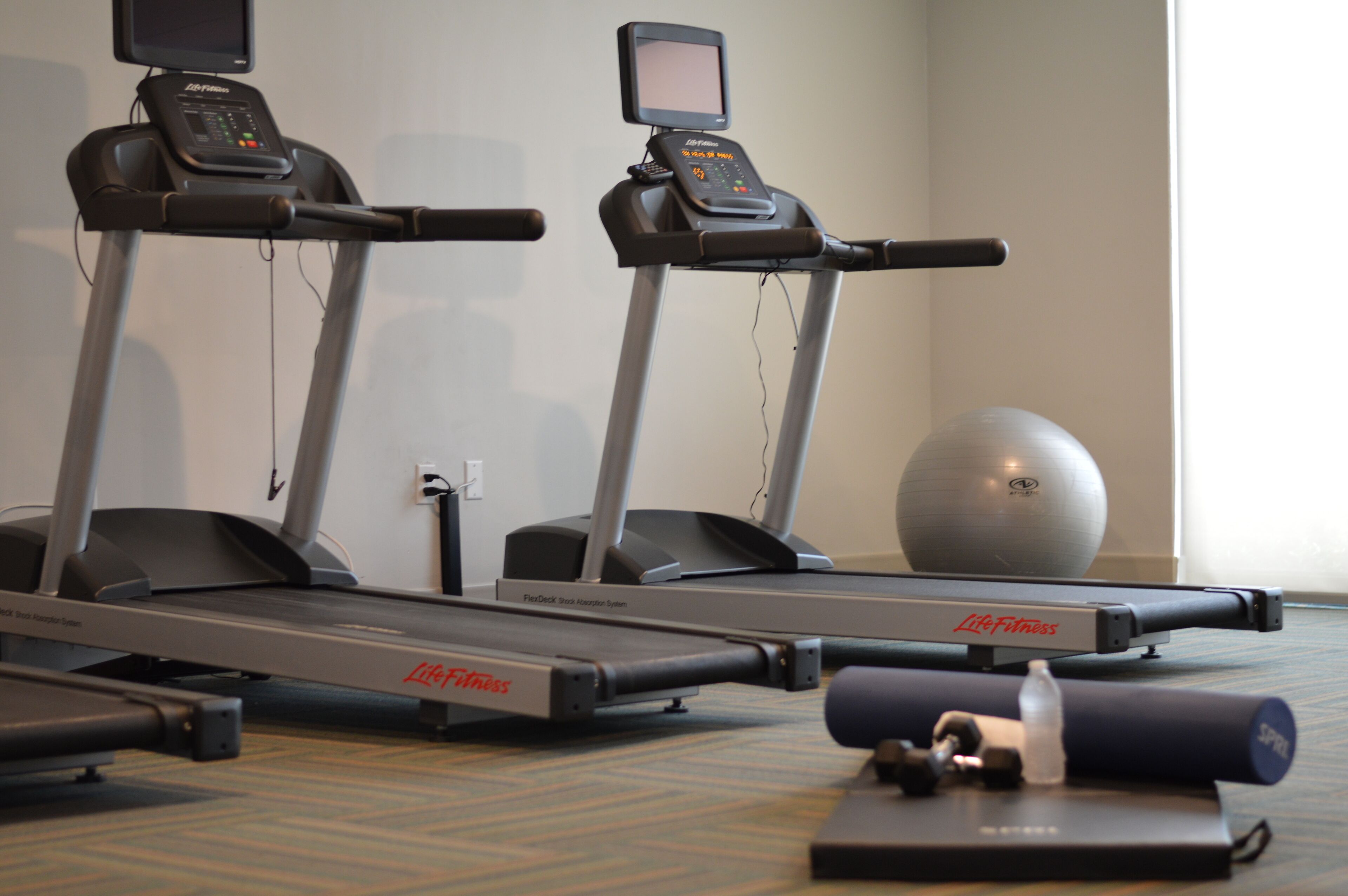 Dos cintas de correr con pantallas digitales, una bicicleta estática y una pelota de ejercicio grande en un gimnasio.