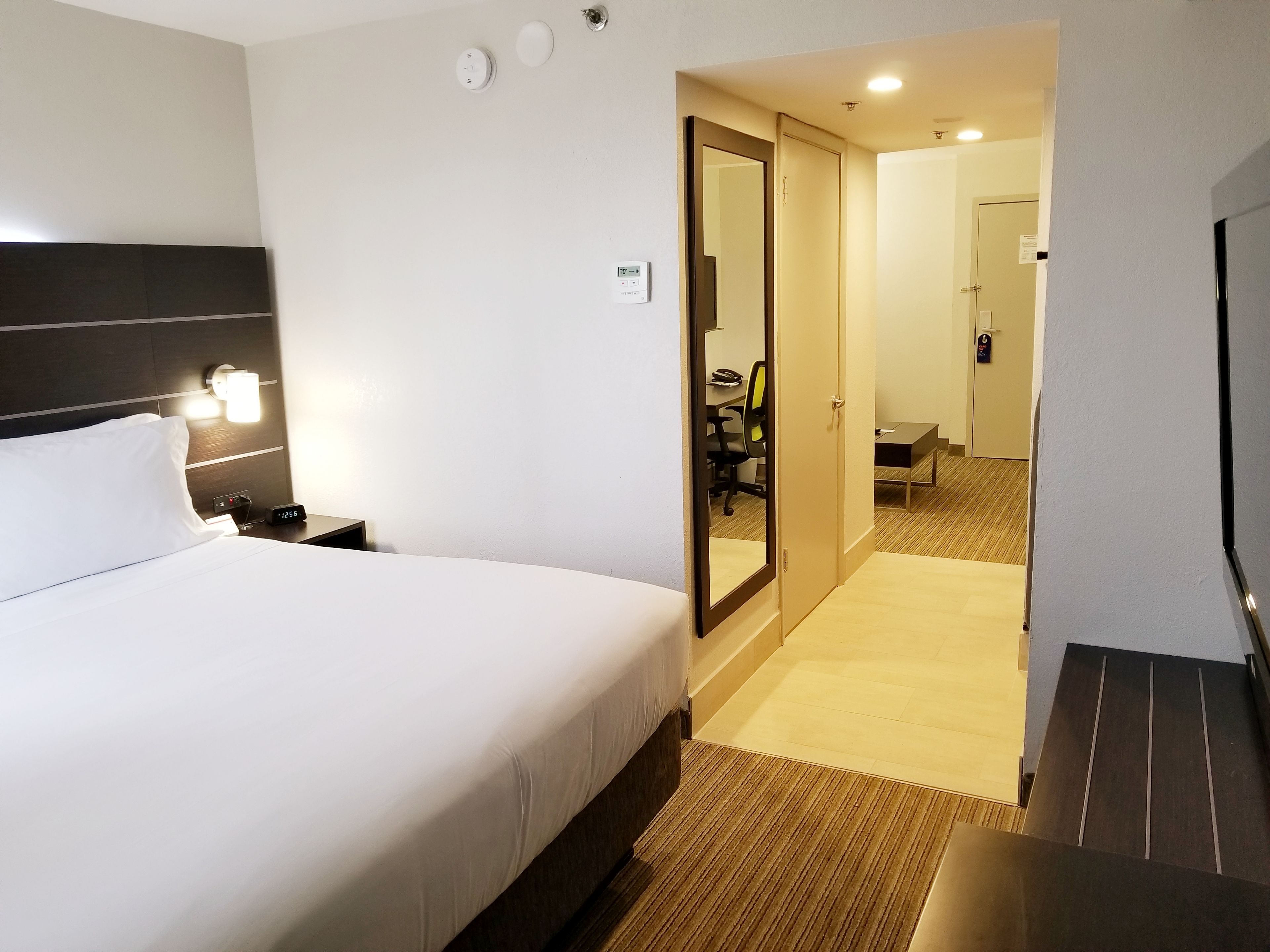 Une chambre d’hôtel avec un lit, une table de chevet, une lampe et un miroir. On y trouve une porte donnant accès à une salle de bain et à un couloir.