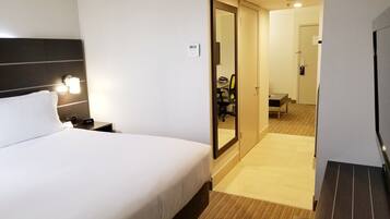 Une chambre d’hôtel avec un lit, une table de chevet, une lampe et un miroir. On y trouve une porte donnant accès à une salle de bain et à un couloir.