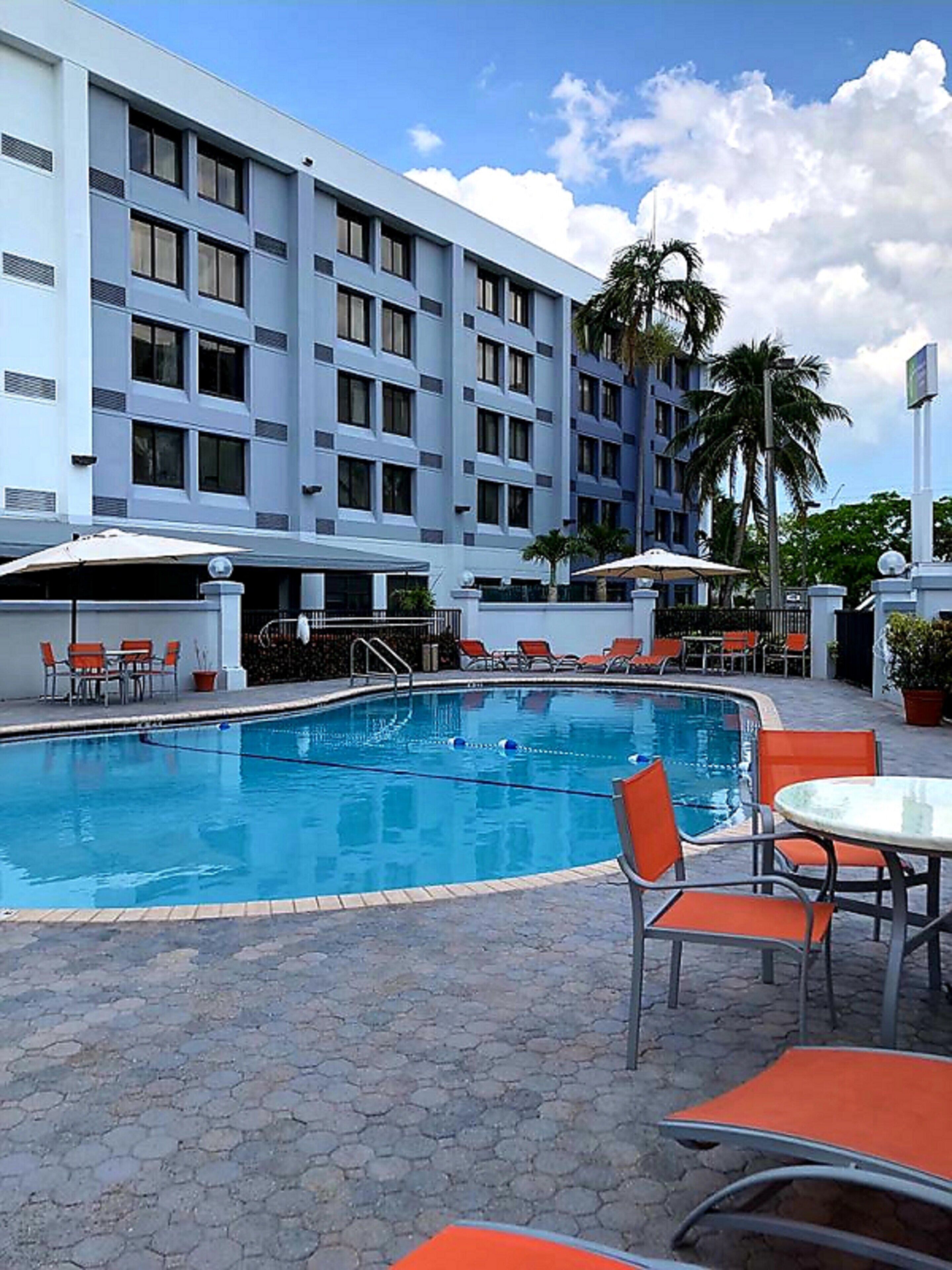 A swimming pool with lounge chairs and umbrellas, surrounded by a hotel building with multiple floors and balconies.
