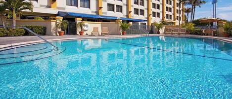A hotel pool with clear blue water, surrounded by palm trees and a building with multiple windows.